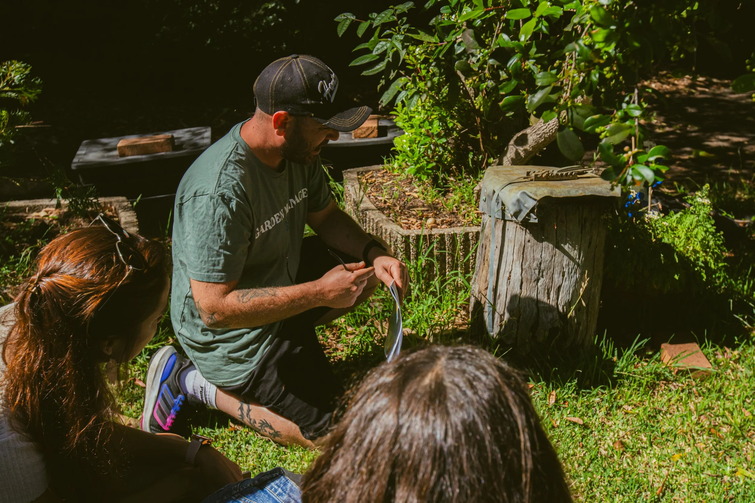 A man kneeling outdoors, holding a pen and notebook, with three women sitting nearby listening. They are in a garden setting with greenery and a tree stump nearby.