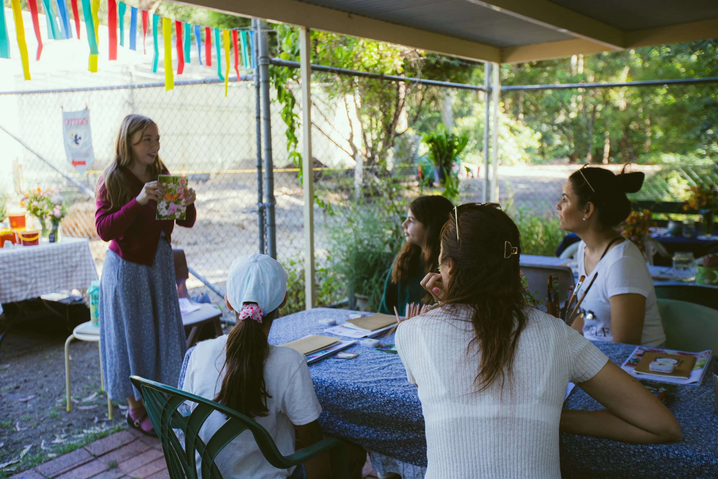 Gemma standing and reading a card to a seated group of women and a girl at an outdoor gathering, with colorful decorations hanging overhead and a table with flowers and supplies in the background.