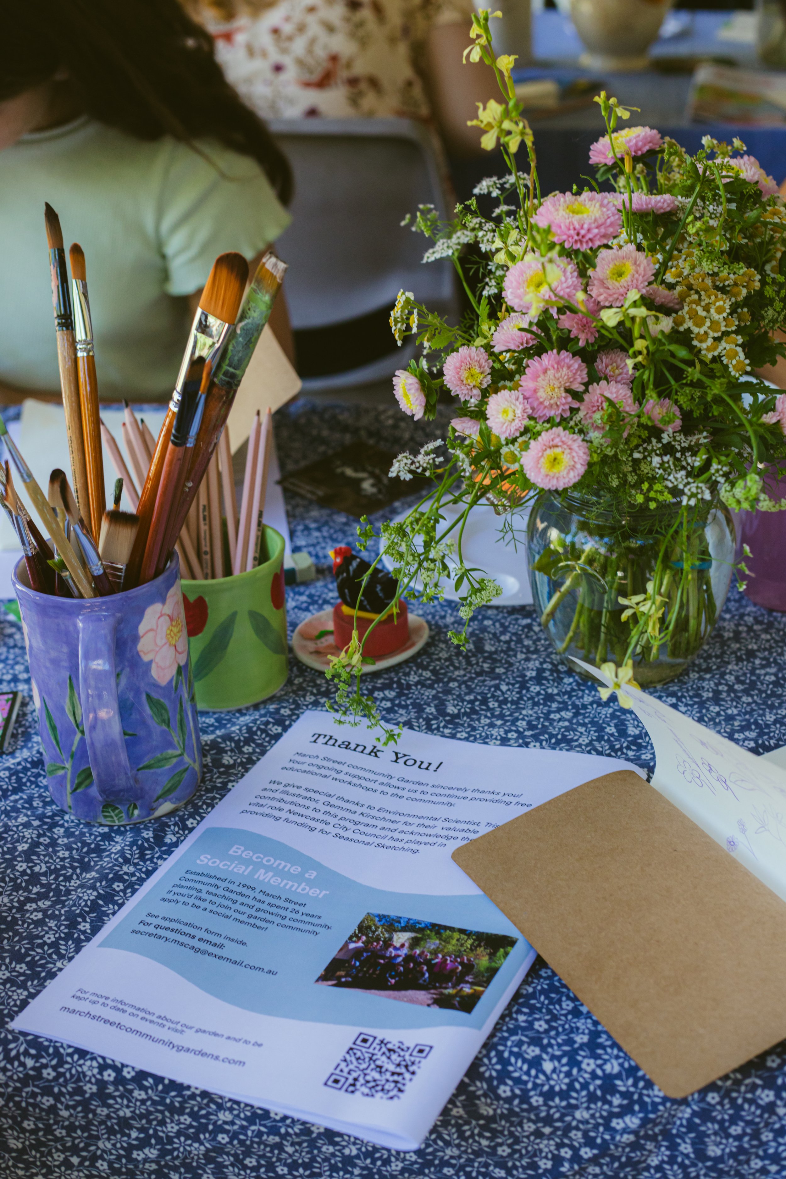 A table decorated with a bouquet of pink and white flowers in a glass jar, a purple painted mug holding paintbrushes, a green container with red hearts, a small decorative black dog figurine on a dish, a brown envelope, and a flyer about becoming a s