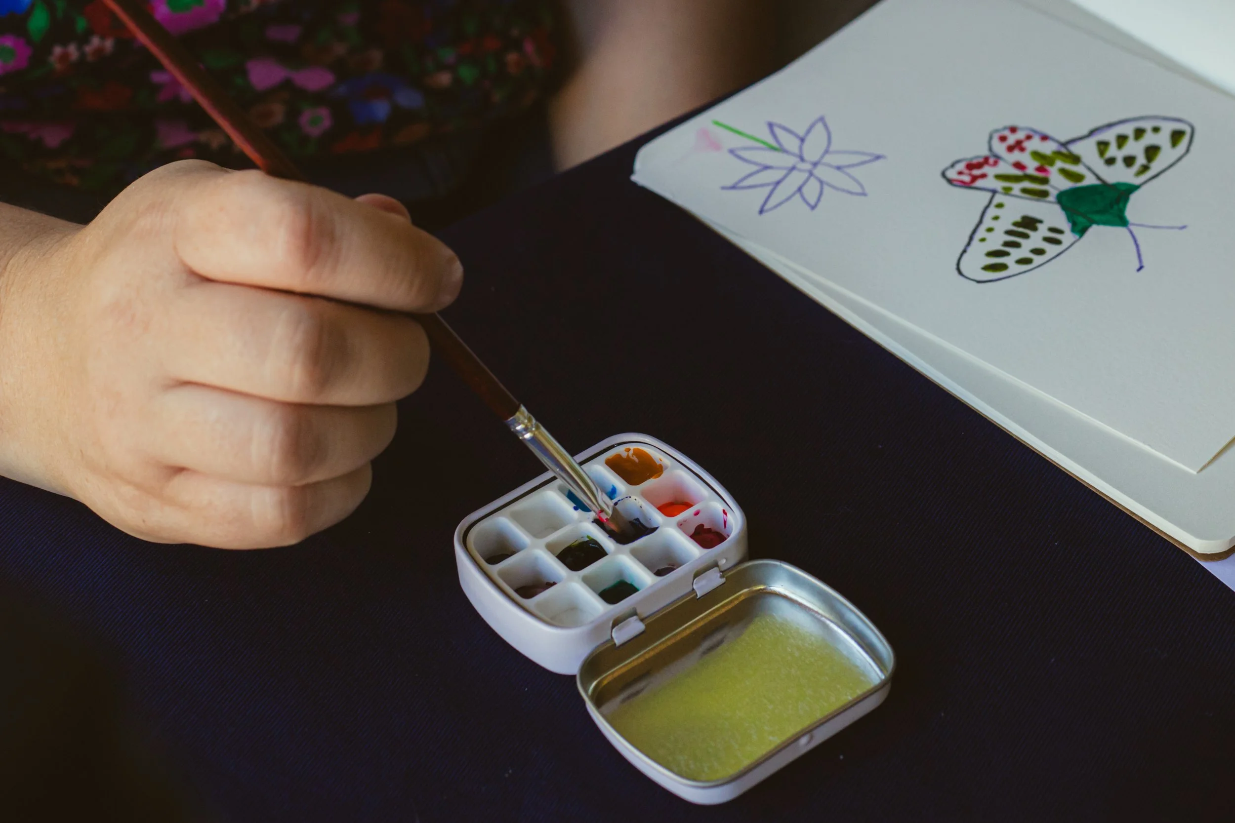 A person painting with watercolors on paper, with a butterfly and flower drawing visible, using a small watercolor palette and brush.