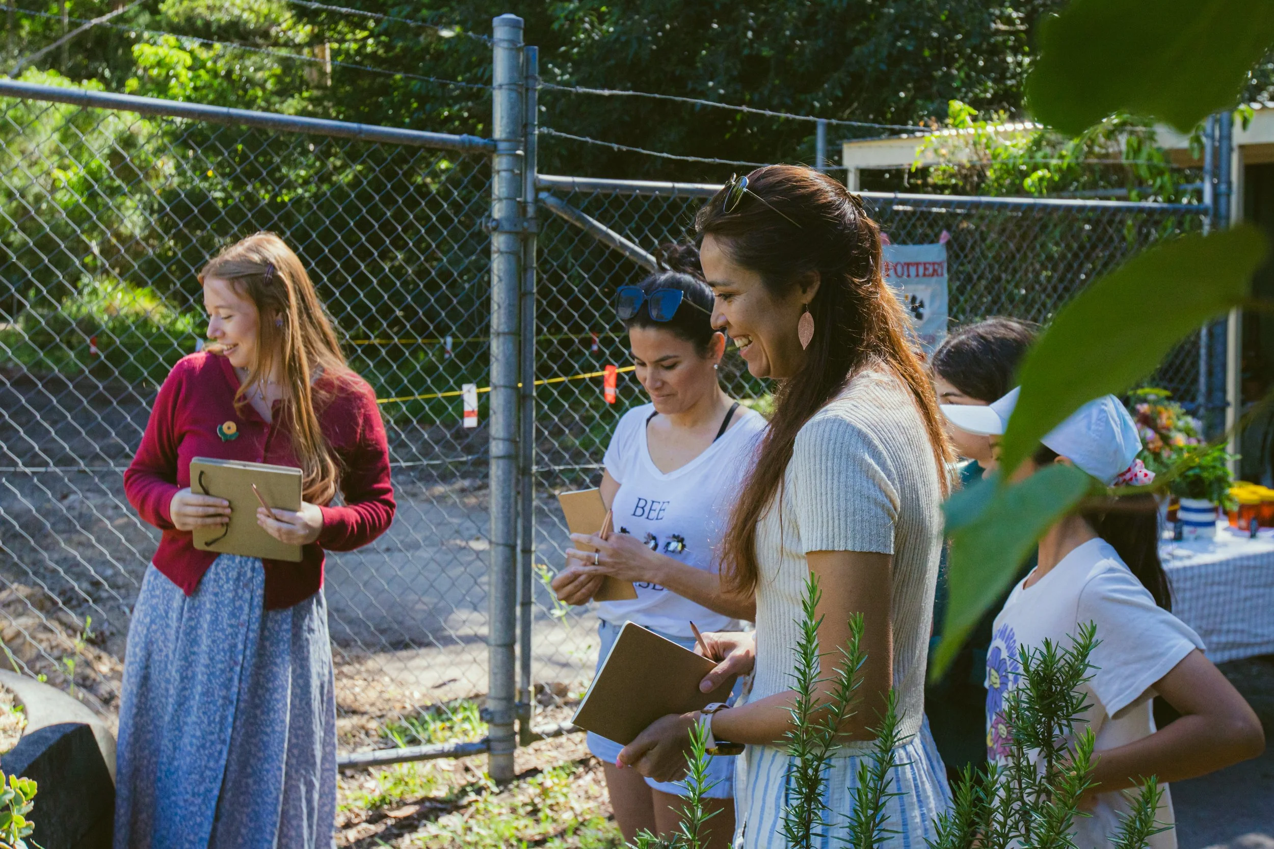 Group of women and girls standing outdoors near a fence, holding notebooks and smiling, possibly participating in an art event.