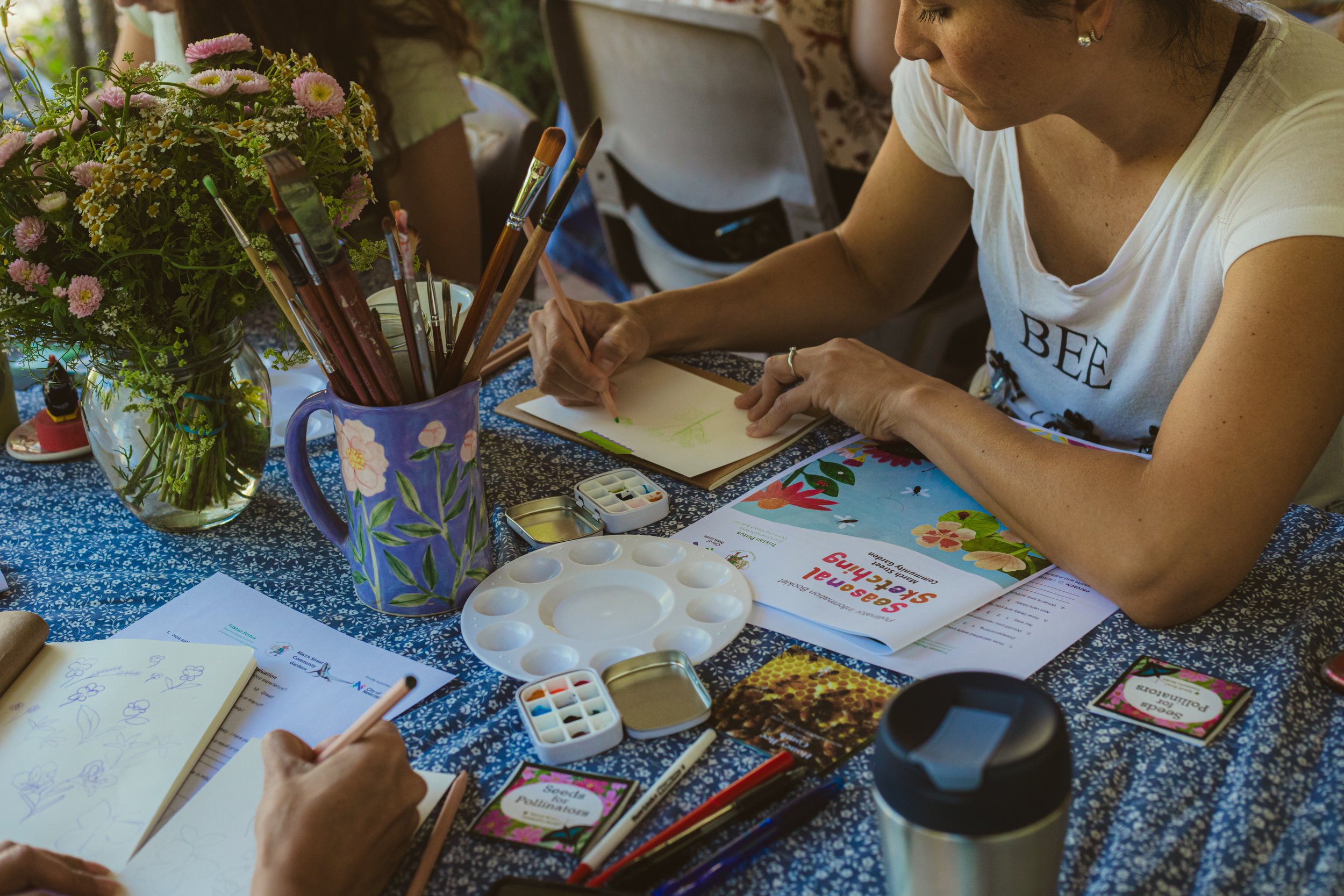 Women painting and drawing at a table with flowers, art supplies, and informational booklets.