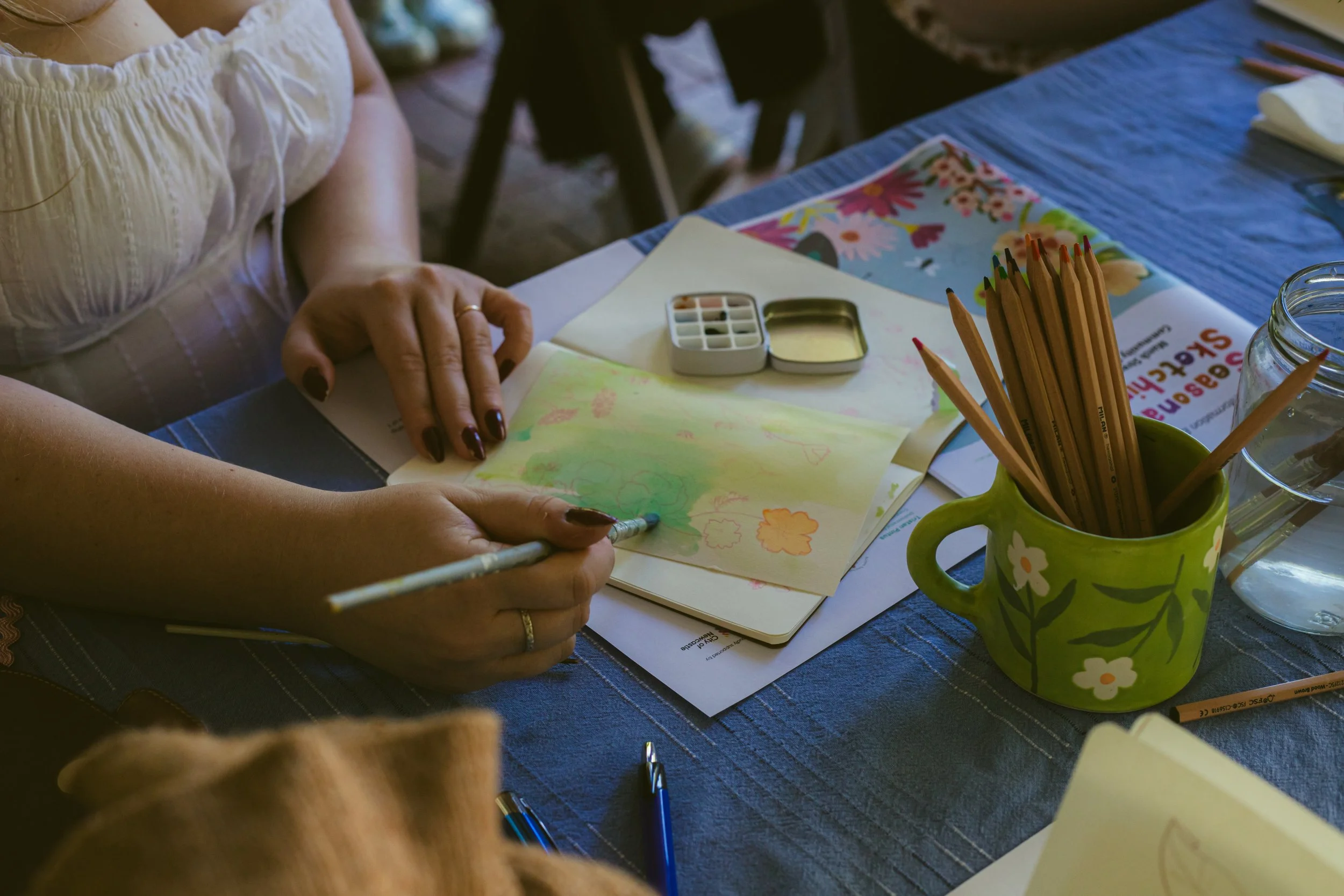 Close-up of a person's hands drawing on paper with colored pencils and watercolor paints, surrounded by art supplies on a blue tablecloth.