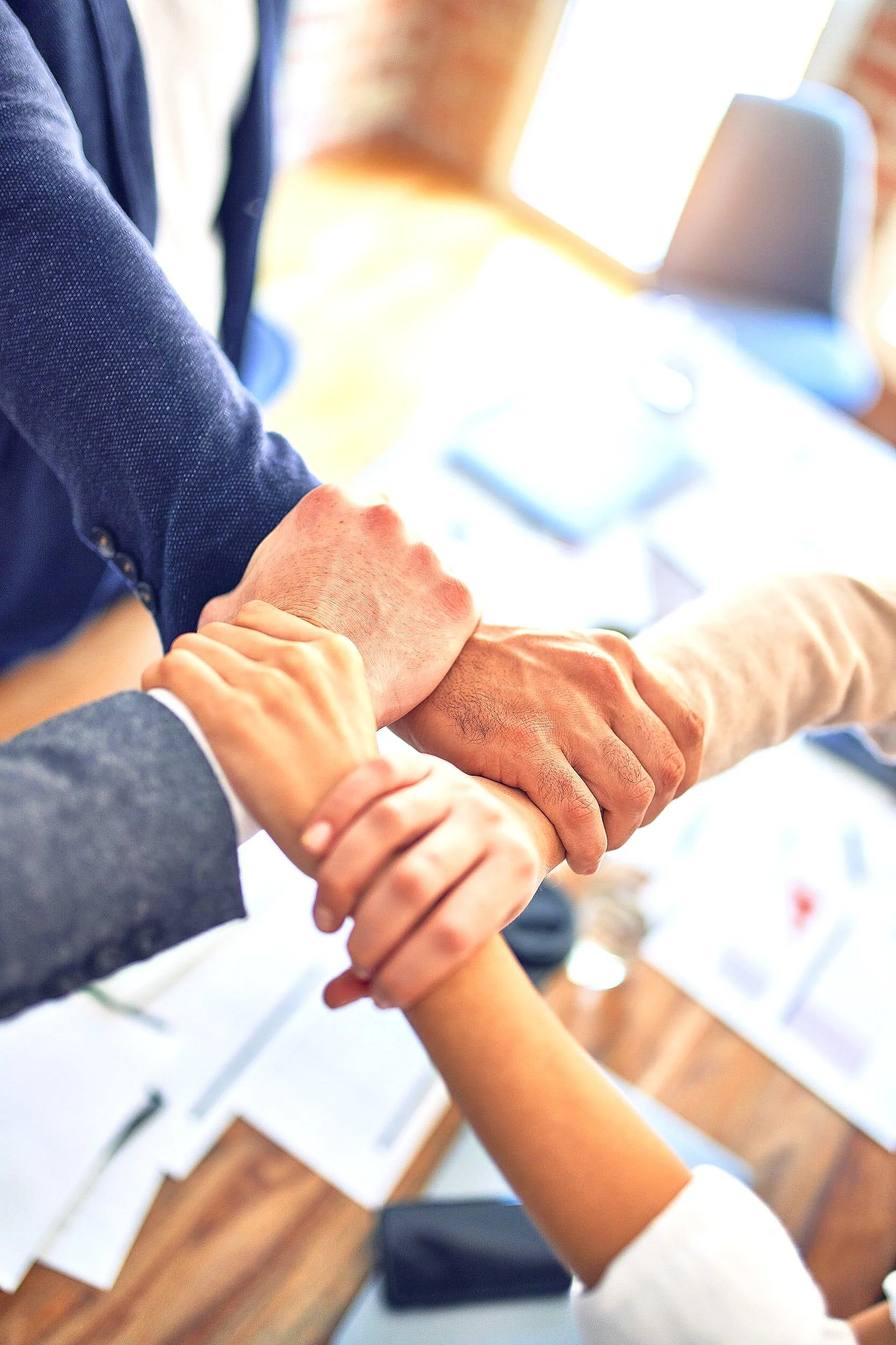 Close-up of four people joining hands in a circle over a wooden table, symbolizing teamwork and unity in an office setting.