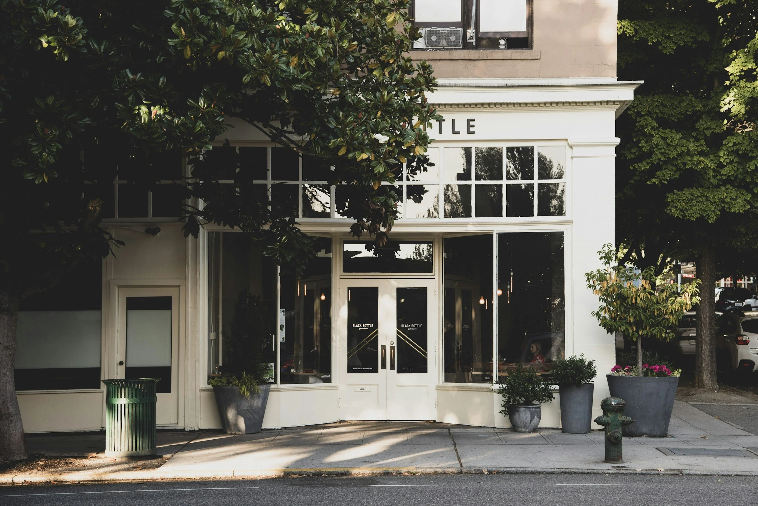 Front view of a storefront with large glass windows and doors, surrounded by potted plants and trees on the sidewalk, with a small green trash can and a fire hydrant nearby.
