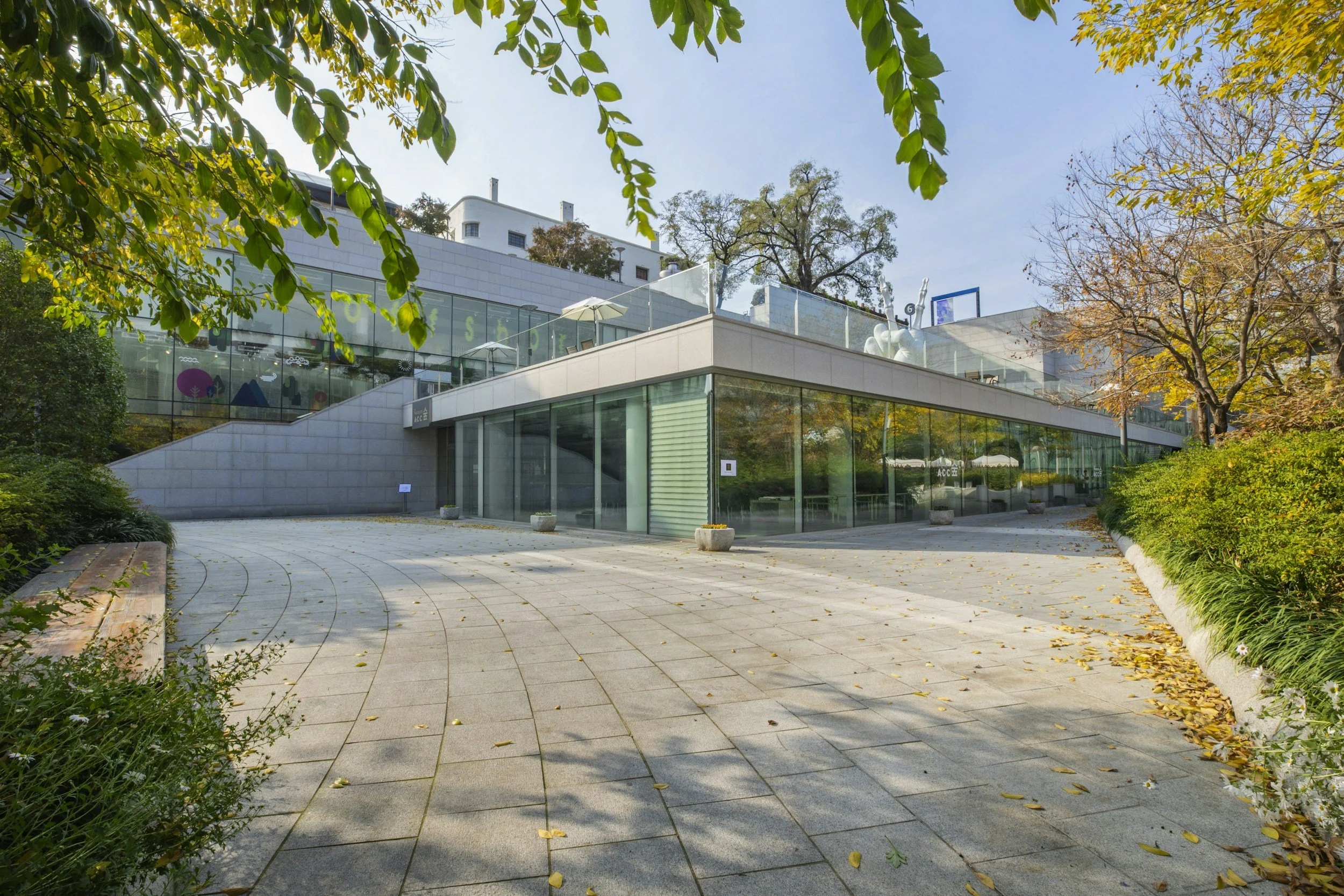 Modern building with large glass windows, surrounded by trees with fall foliage, and a paved walkway in front.