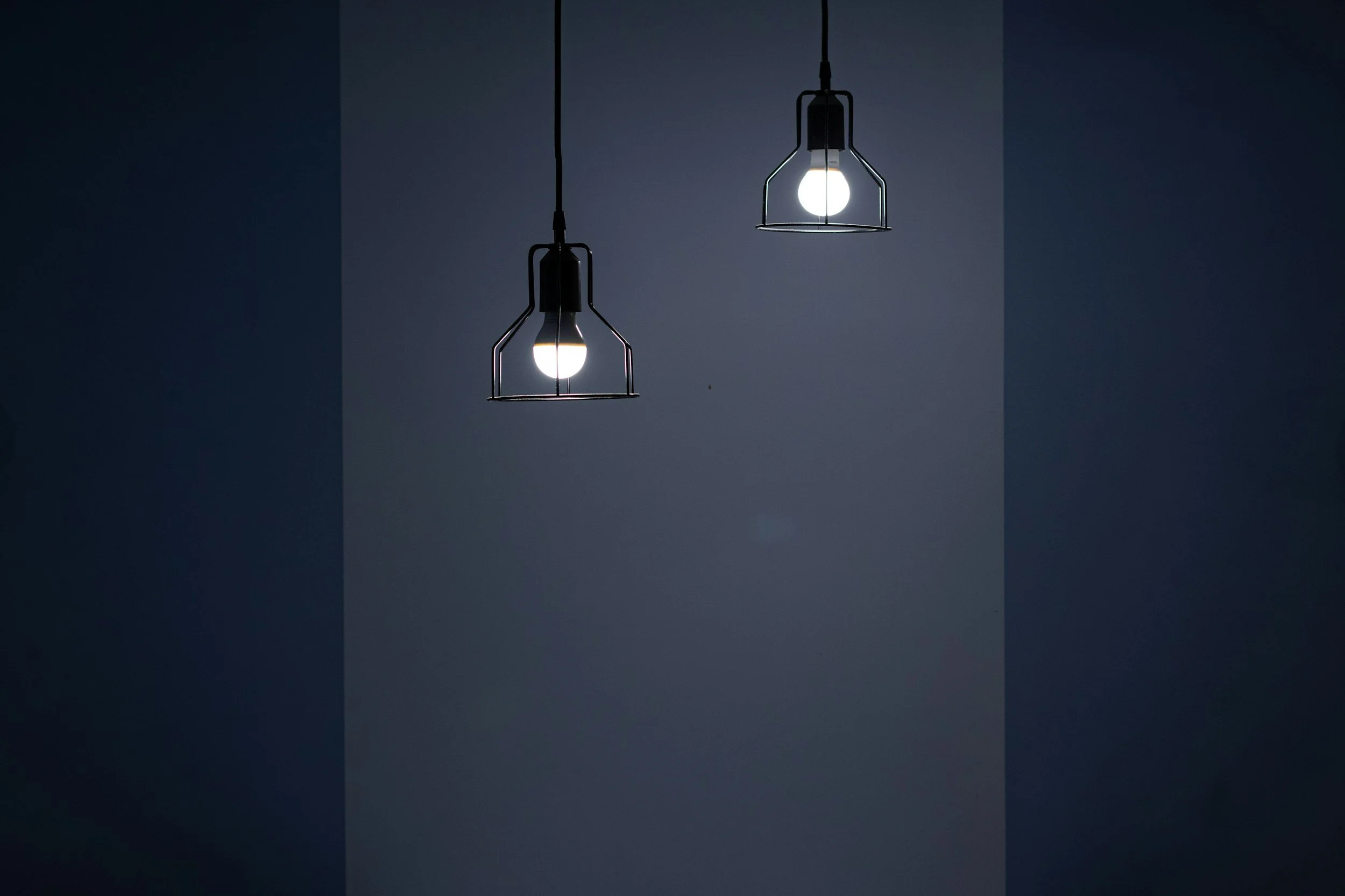 Two hanging light bulbs with wire cages illuminate against a two-tone wall, one light bulb is turned on and the other is off.