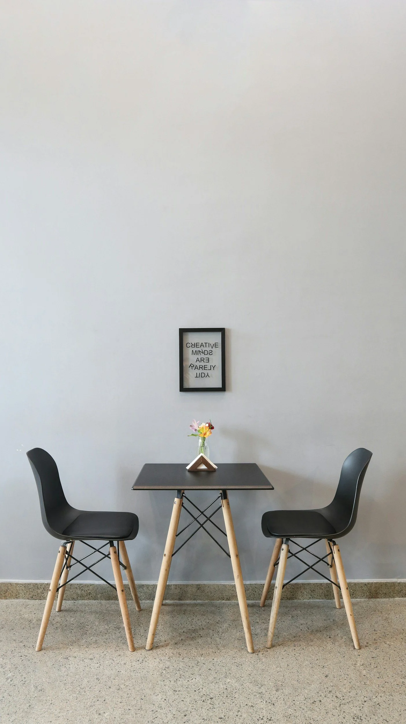 A small black table with two black chairs with wooden legs, a glass vase with pink and white flowers, and a triangular object on the table. There is a framed word art hung on a gray wall behind the table that reads 'CREATIVE MINDSET ARE RARELY ALONE'.