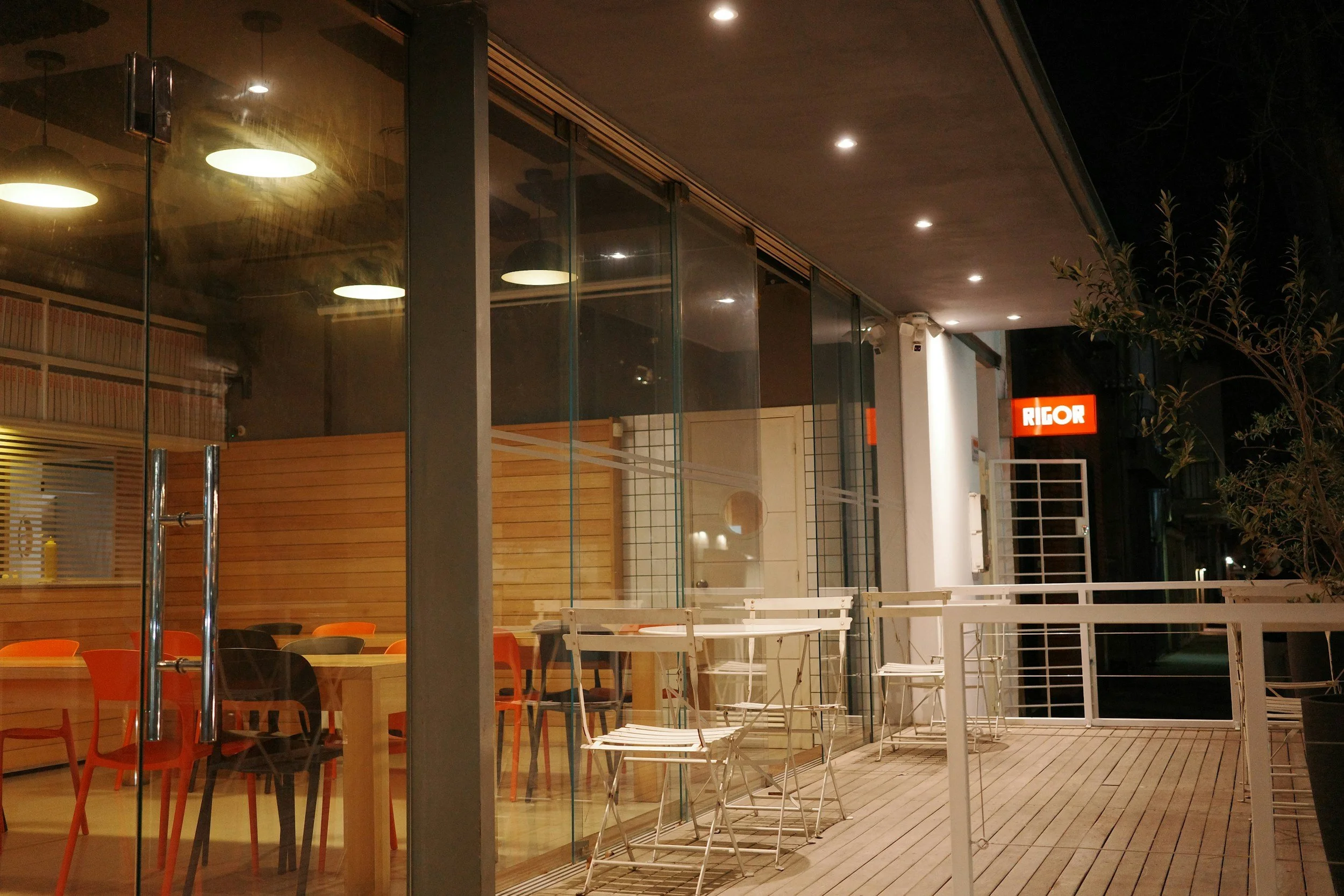 Empty outdoor patio at a restaurant or café at night, with glass walls, white tables and chairs, and a building with a red Rigor sign illuminated in the background.