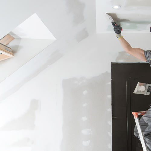 Person using a trowel to plaster the ceiling in a room under renovation, with drywall and a window nearby.
