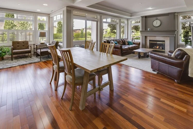 Bright living and dining room with hardwood floors, large windows, a wooden dining table with chairs, a leather sofa, a fireplace, and a clock on the mantel.