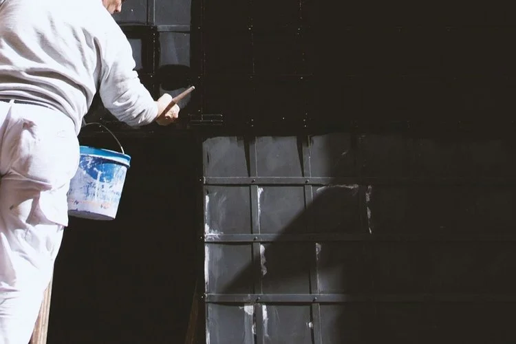 Person wearing white clothing holding a bucket and a tool, working on a black wall with panels.