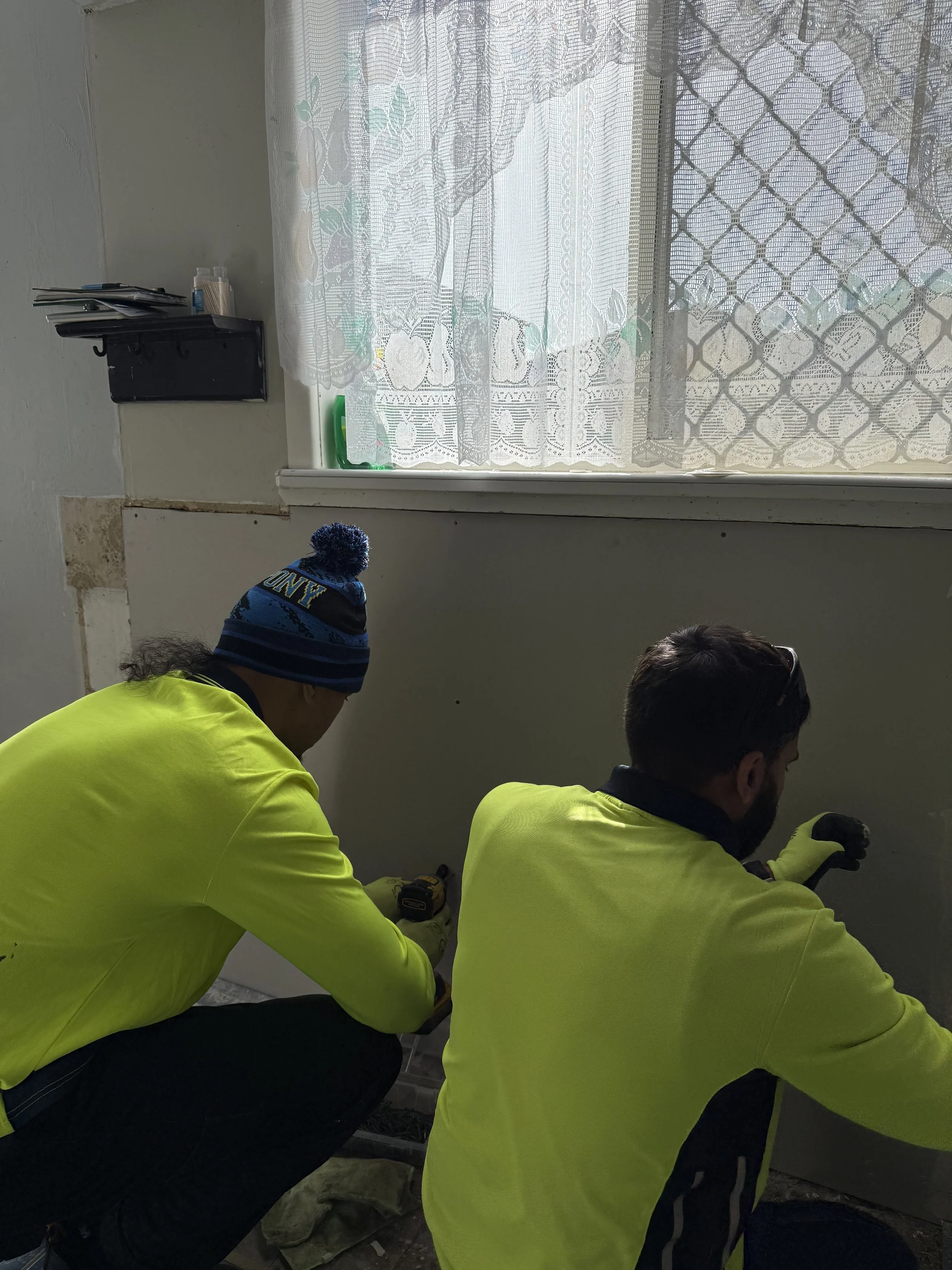 Two workers in bright yellow jackets working on a wall near a window with lace curtains. One worker is wearing a blue knit hat with 