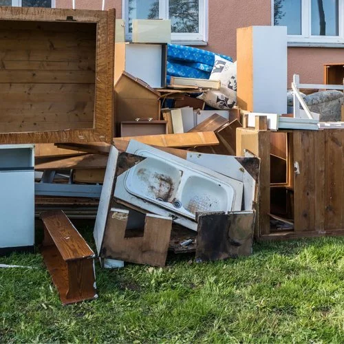 Piles of discarded furniture and household items outside a house, including a broken kitchen sink, wooden cabinets, a small bench, and various boxes and materials.