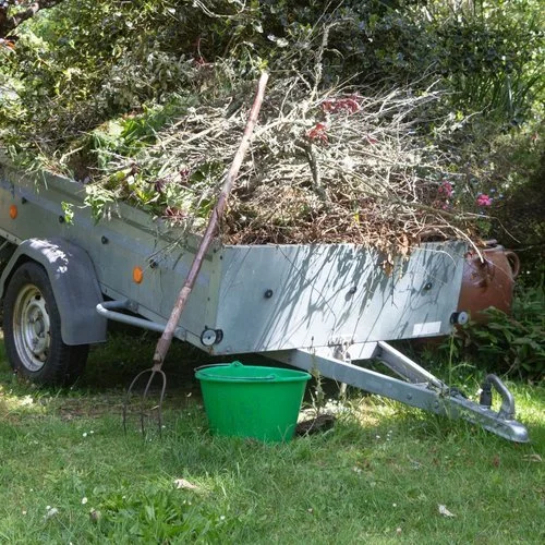 A yard trailer filled with yard debris, a rake leaning against it, and a green bucket on the grass next to it.