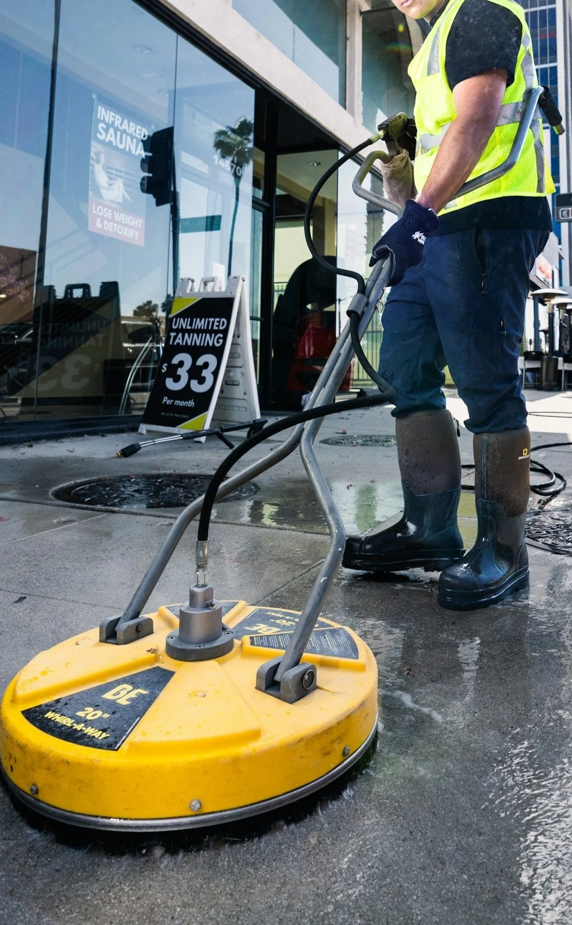 A person wearing black boots, blue pants, gloves, and a yellow safety vest uses a yellow power washer to clean a wet sidewalk outside a storefront with signs for infrared sauna, tanning, and weight loss.