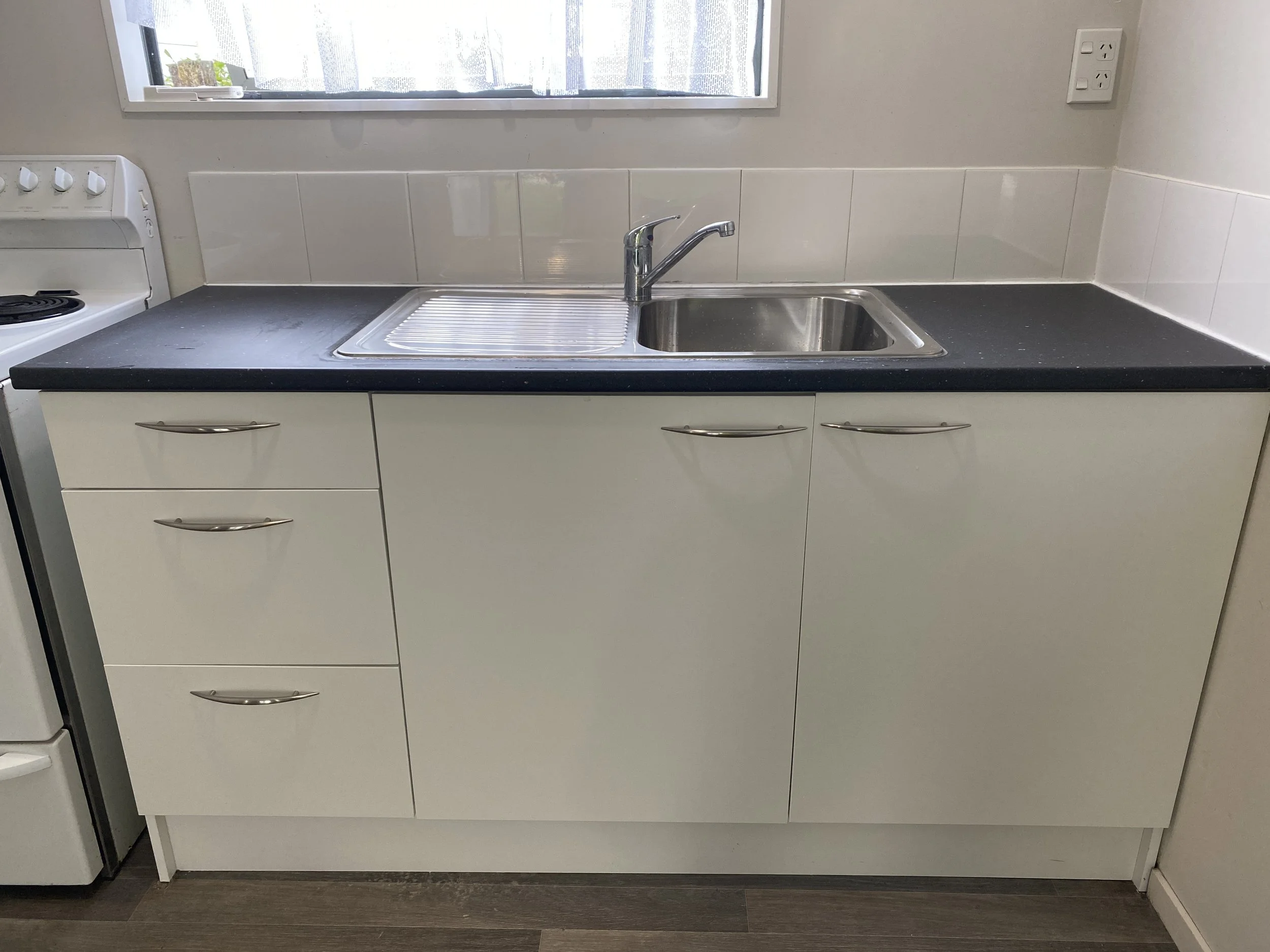 Kitchen sink with faucet, black countertop, white cabinets, white tiled backsplash, window above sink, electrical outlet on wall.