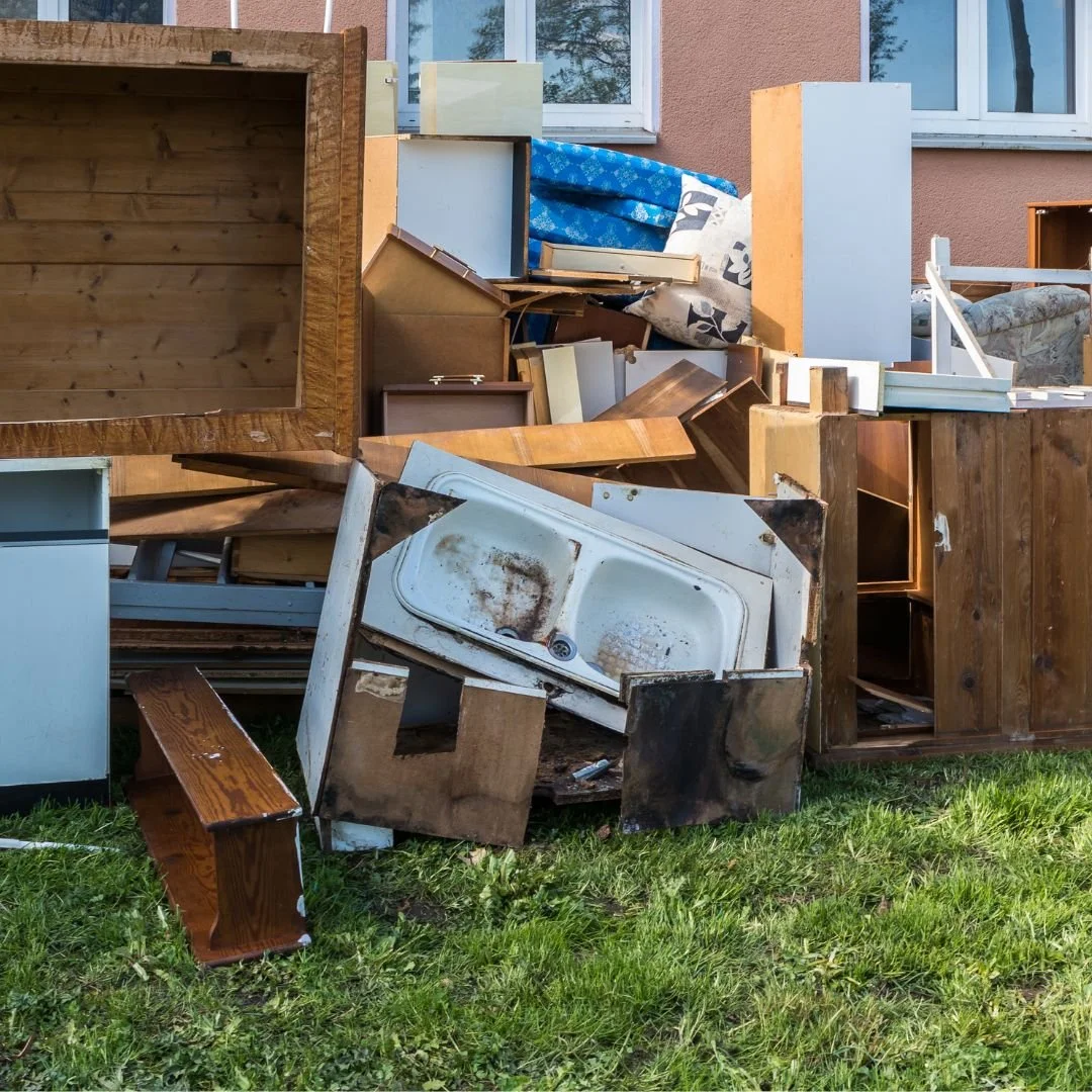 Pile of discarded furniture and household items including broken cabinets, a rusty sink, and wooden furniture parts outdoors on grass.