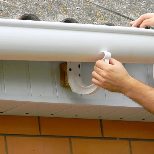 A person installing a white gutter on a house with brick siding, using a hand to hold the gutter in place.