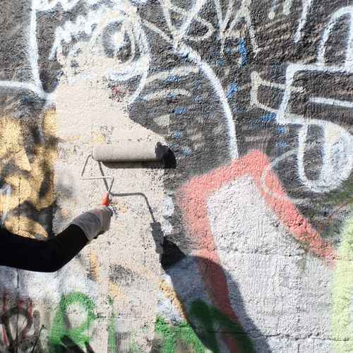 Person using paint roller on graffiti-covered wall.