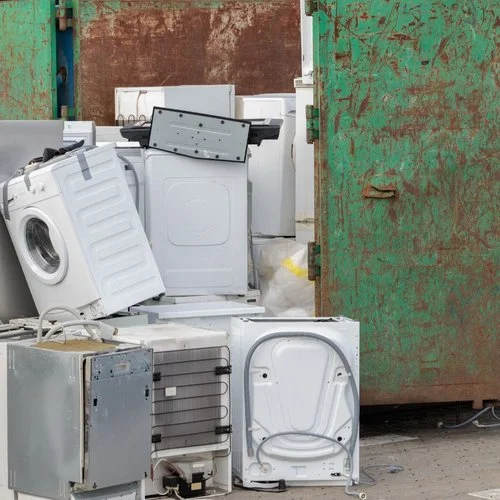 Pile of discarded washing machines and appliances outside next to a rusty green metal door.