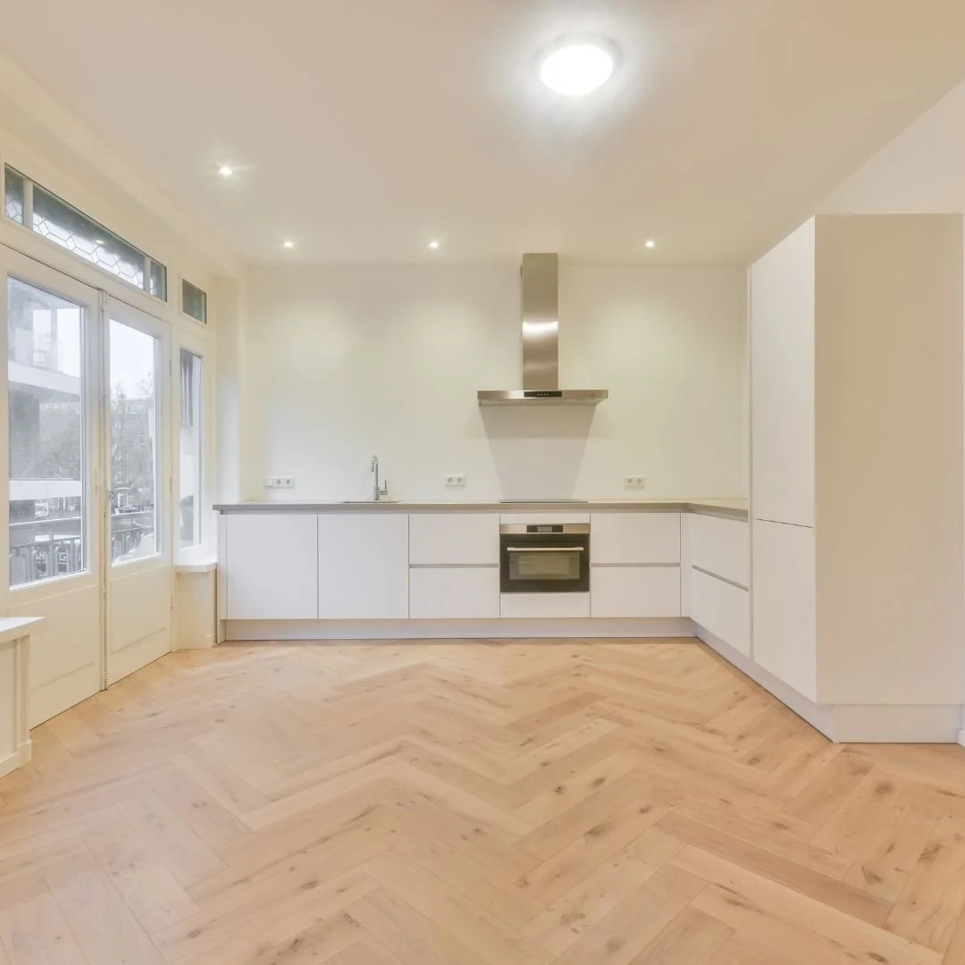 Empty modern kitchen with white cabinets, stainless steel oven and range hood, wooden herringbone floor, and large windows.