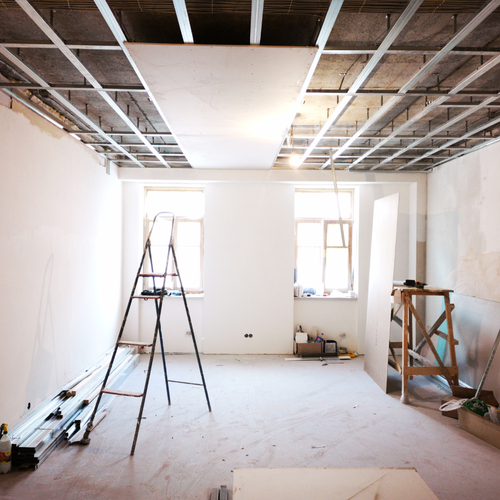 Interior of a room under construction with a stepladder, construction tools, and building materials, with exposed ceiling framework and sunlight coming through windows.