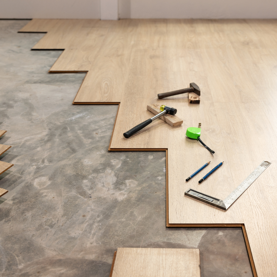 Wood flooring planks and tools including a hammer, measuring tape, level, square, and pencils arranged on a concrete floor, in the process of being installed.