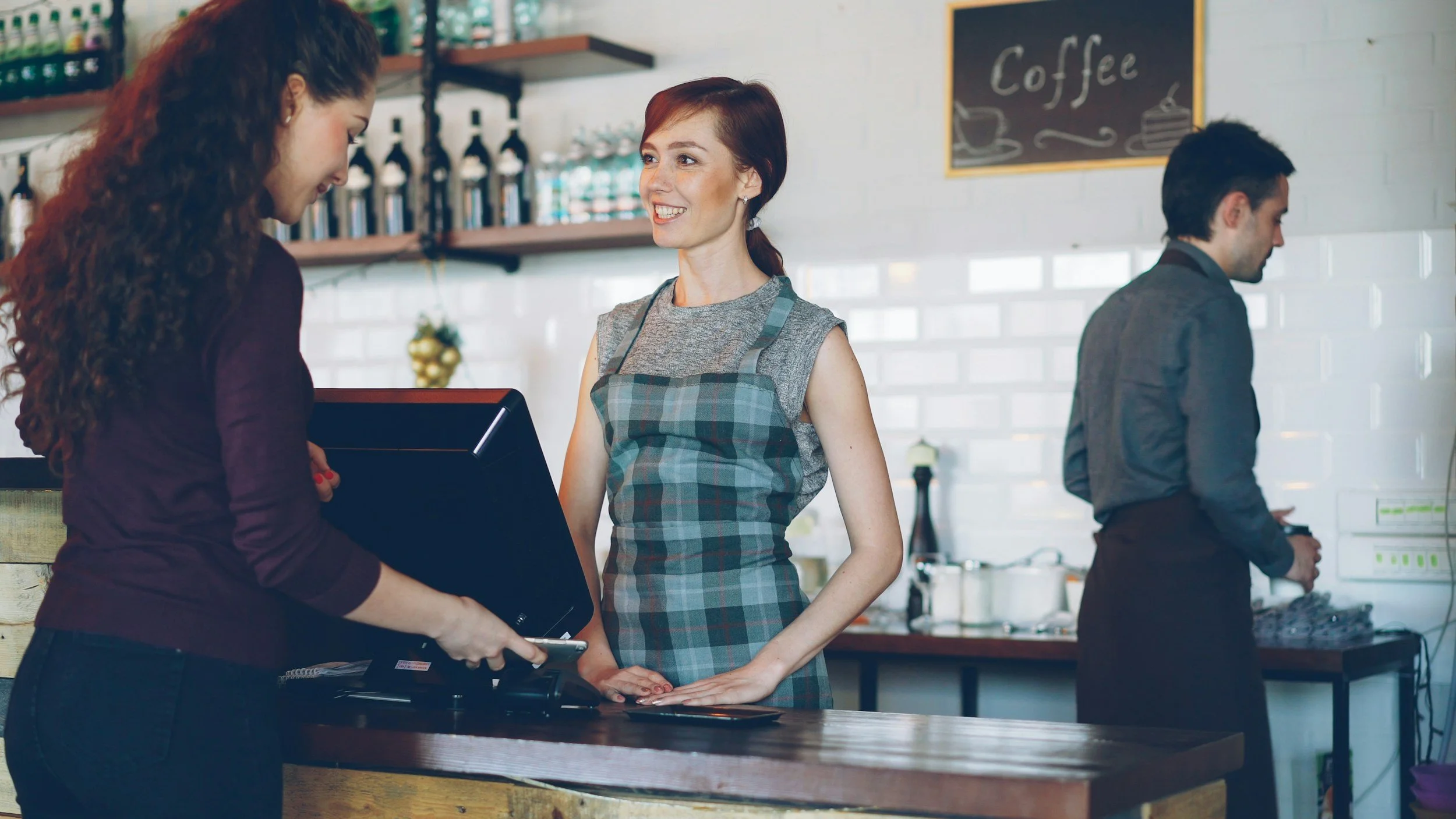 A female in a checked apron serving a customer at a café counter on the Customer Service Practitioner apprenticeship