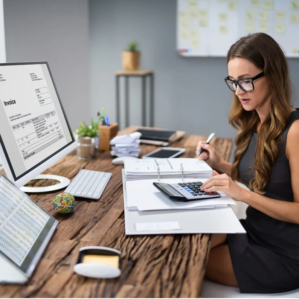 A woman at her computer desk with a large file and calculator is on the Accounts or Finance Apprenticeship