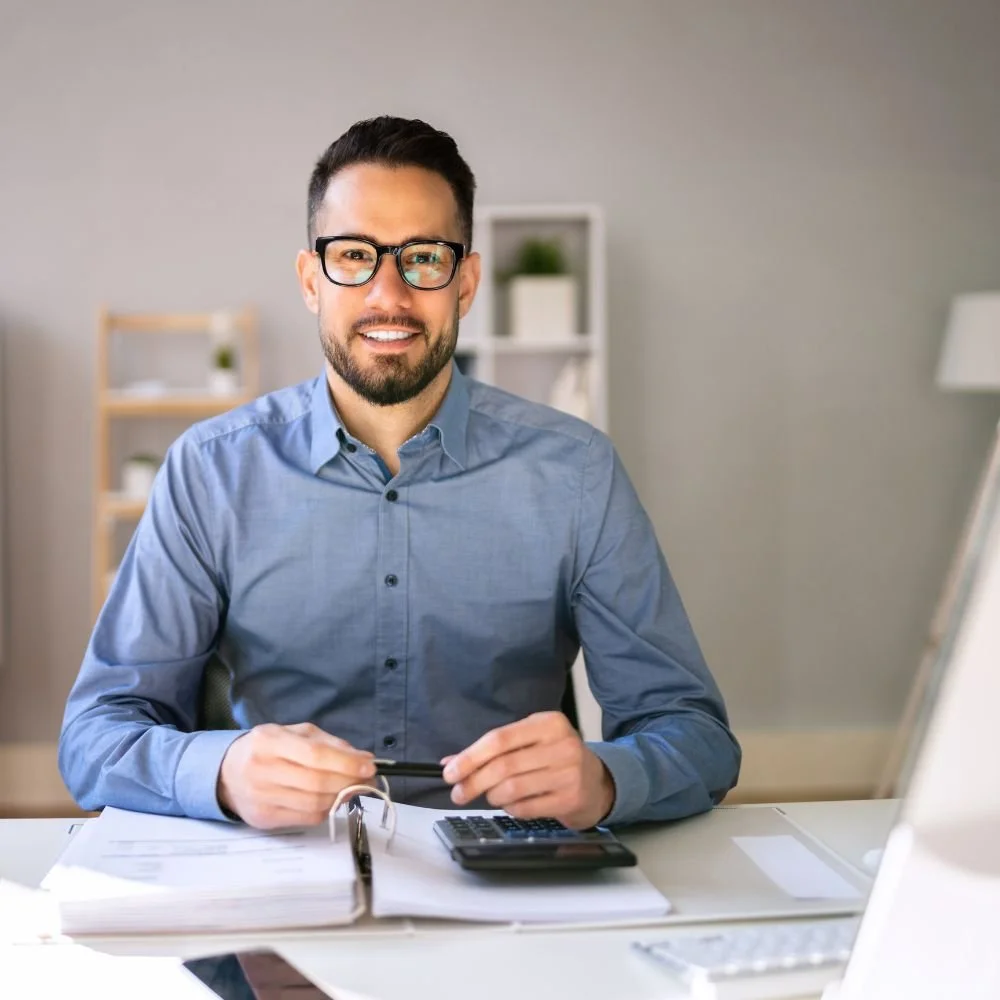 A bearded man in a blue shirt at a desk with a file and calculator in front of hi, during the Assistant accountant apprenticeship