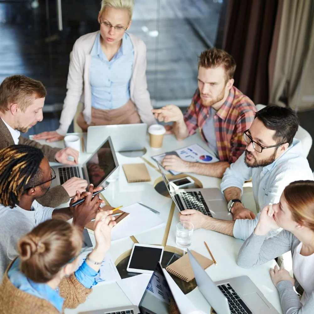 A group of male and female office workers around a desk with their laptops and paperwork, a female at the head of the table is leading the training for employee development