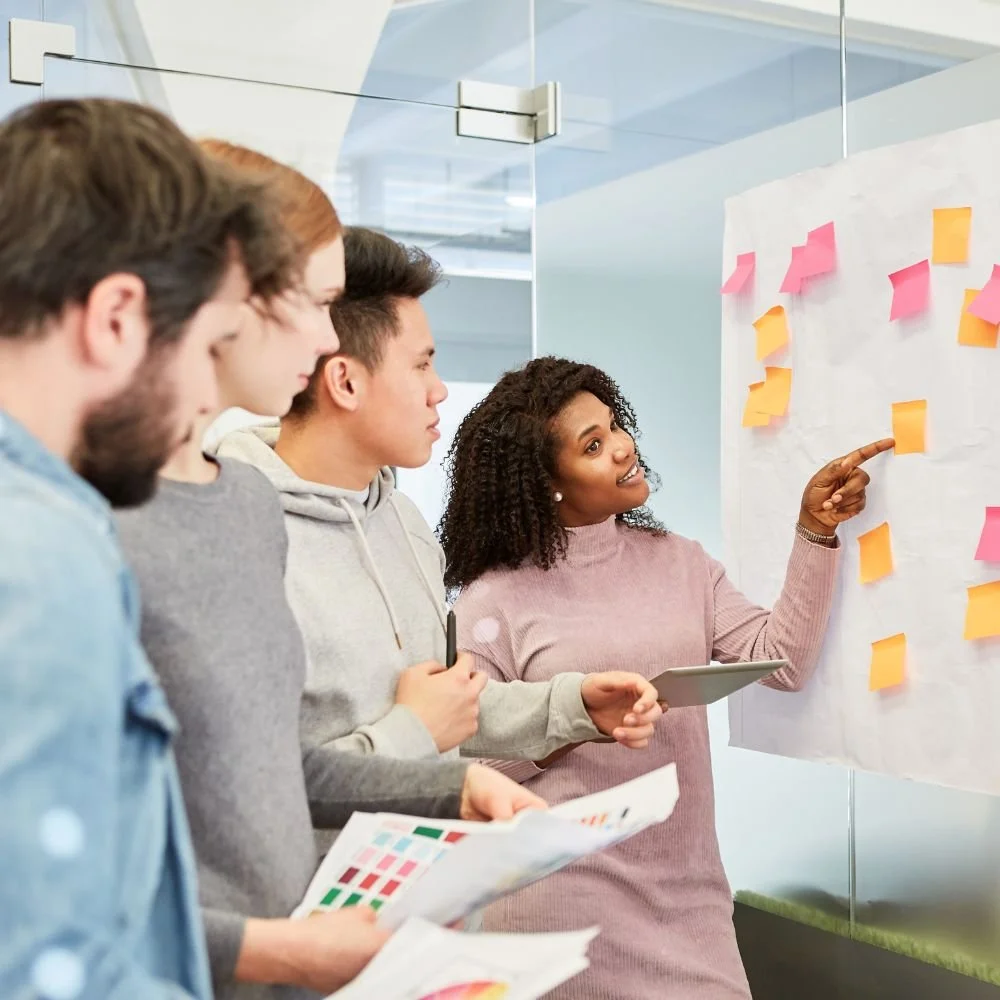 Training for employee development a Black female tutor pointing to post-it notes on a whiteboard to 3 students