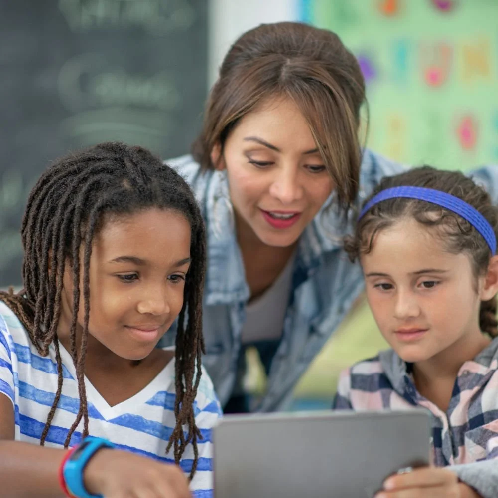 recruit and train professional TAs - A female TA leaning between a boy and a girl working on a laptop in a classroom