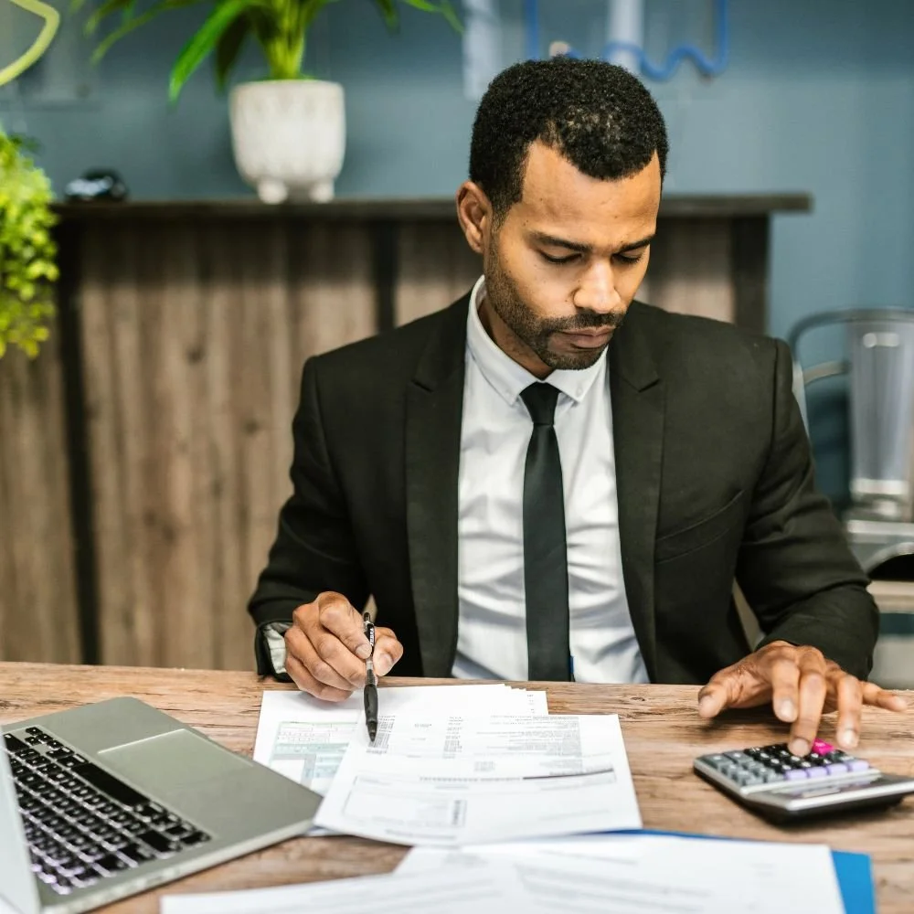 A man in a black suit at a desk with a calculator is on the Accounts or Finance Assistant Apprenticeship