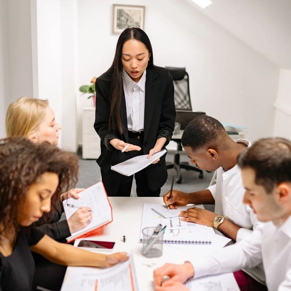 An Asian lady in a black trouser suit is conducting business training with 4 individuals sat as a desk taking notes