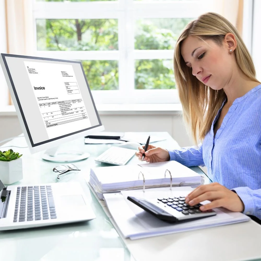 A young lady on an Assistant Accountant Apprenticeship working at her desk with an invoice on her computer, paperwork and a calculator