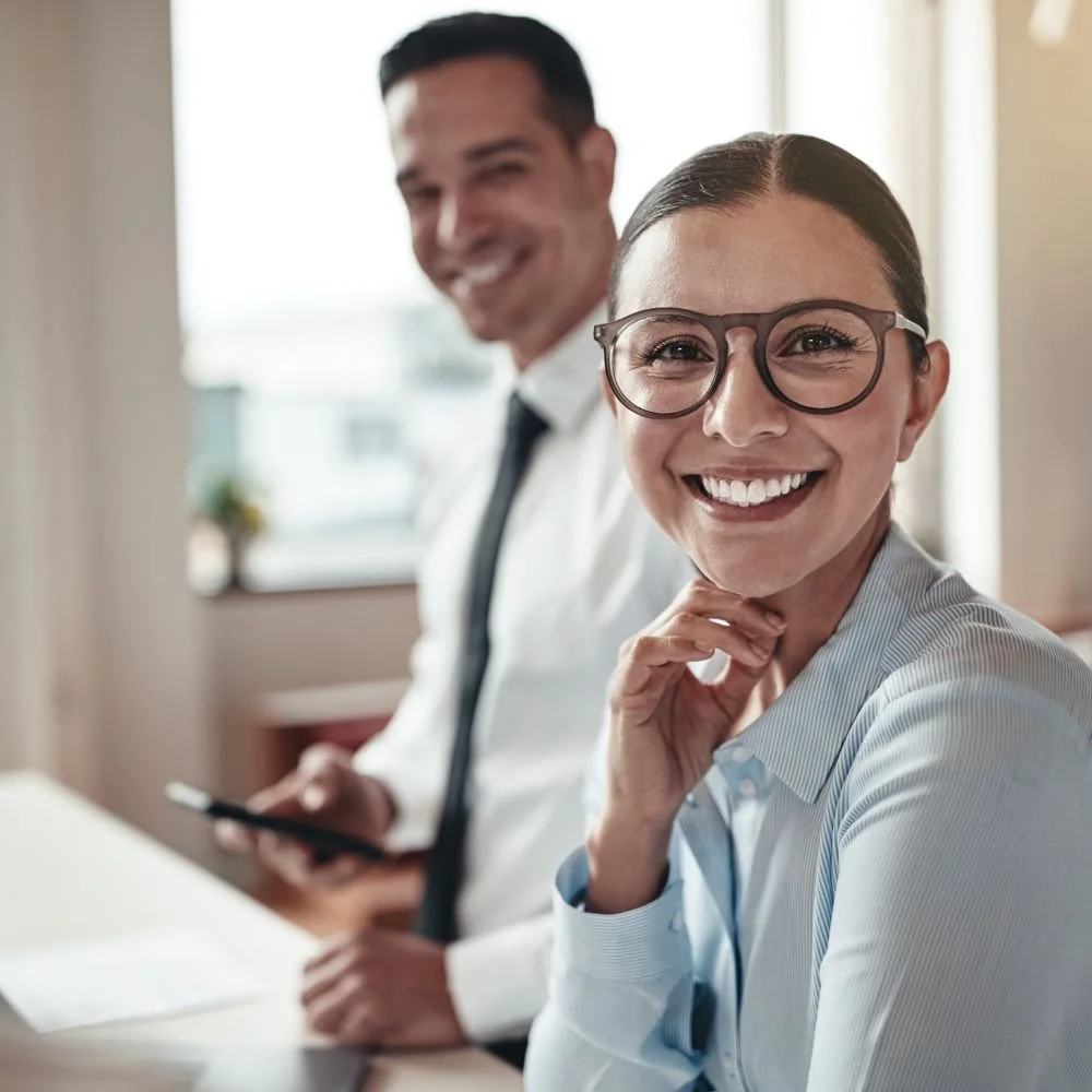 A young female business woman wearing glasses smiling to the camera with a male colleague in the background next to her for off-the-job training hours in apprenticeships