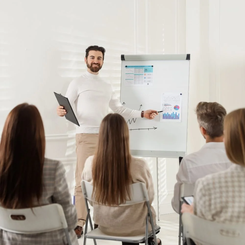 A bearded man pointing to graphs on a flip chart is doing group training for employee development to a mixed group of office workers