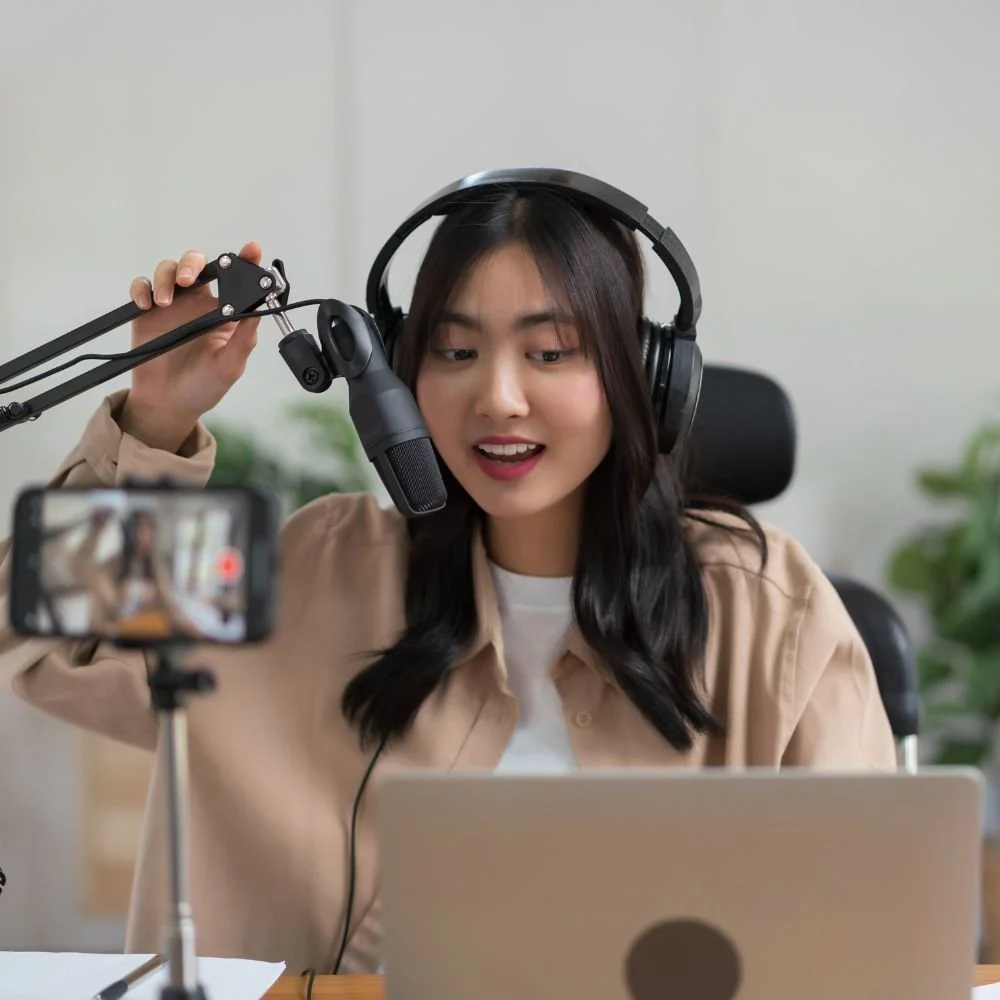 A young Asian lady at a laptop with headphones on and recording equipment during a Content Creator Apprenticeship