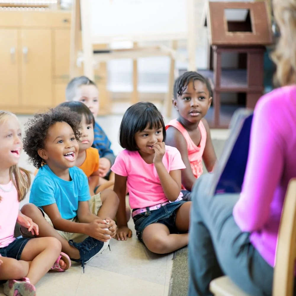 Young children sat on a classroom floor listening to a female on the Teaching assistant apprenticeship read a story