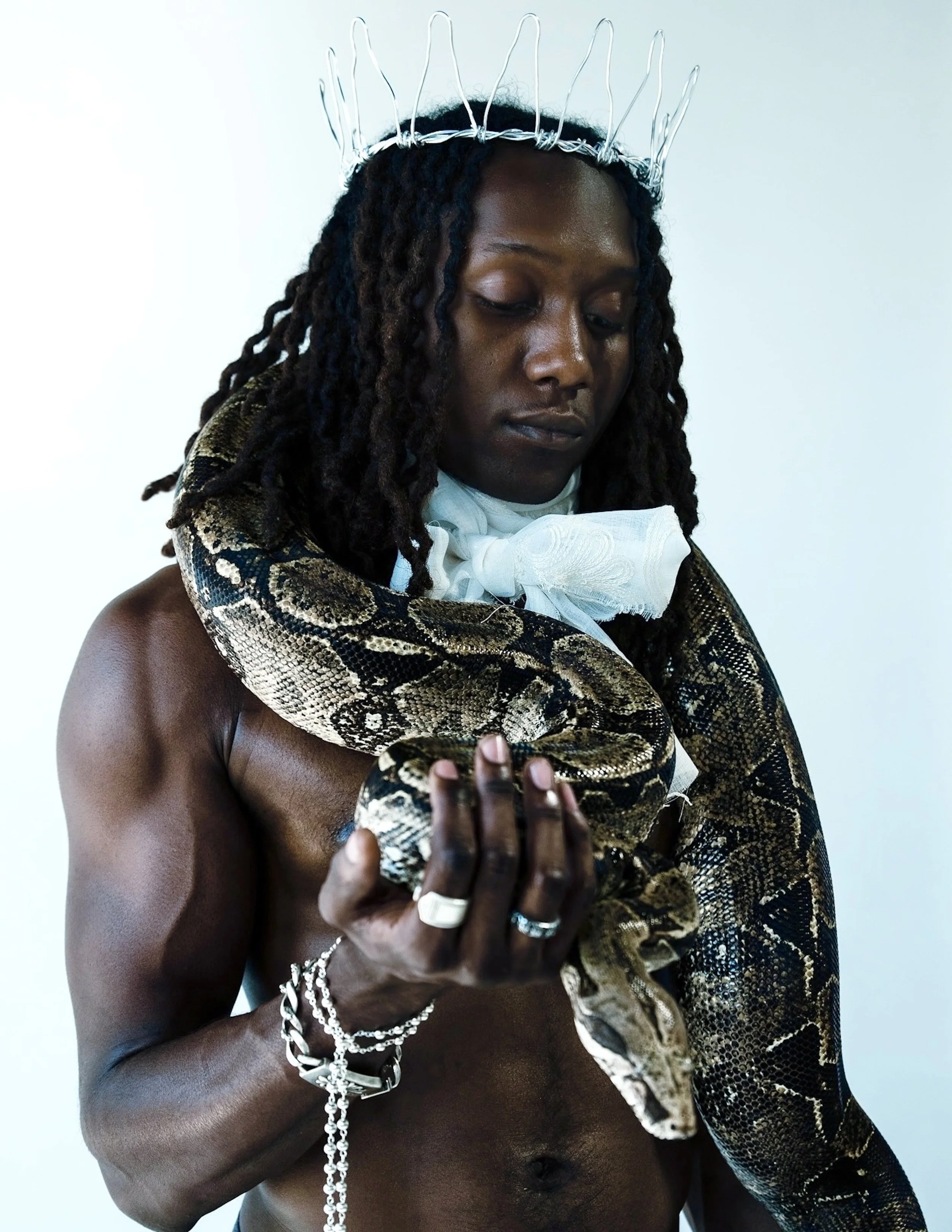 A shirtless man wearing a white tie and a wire crown, holding a large snake draped around his neck and arm, with a plain white background and a rosary hanging from his hand.