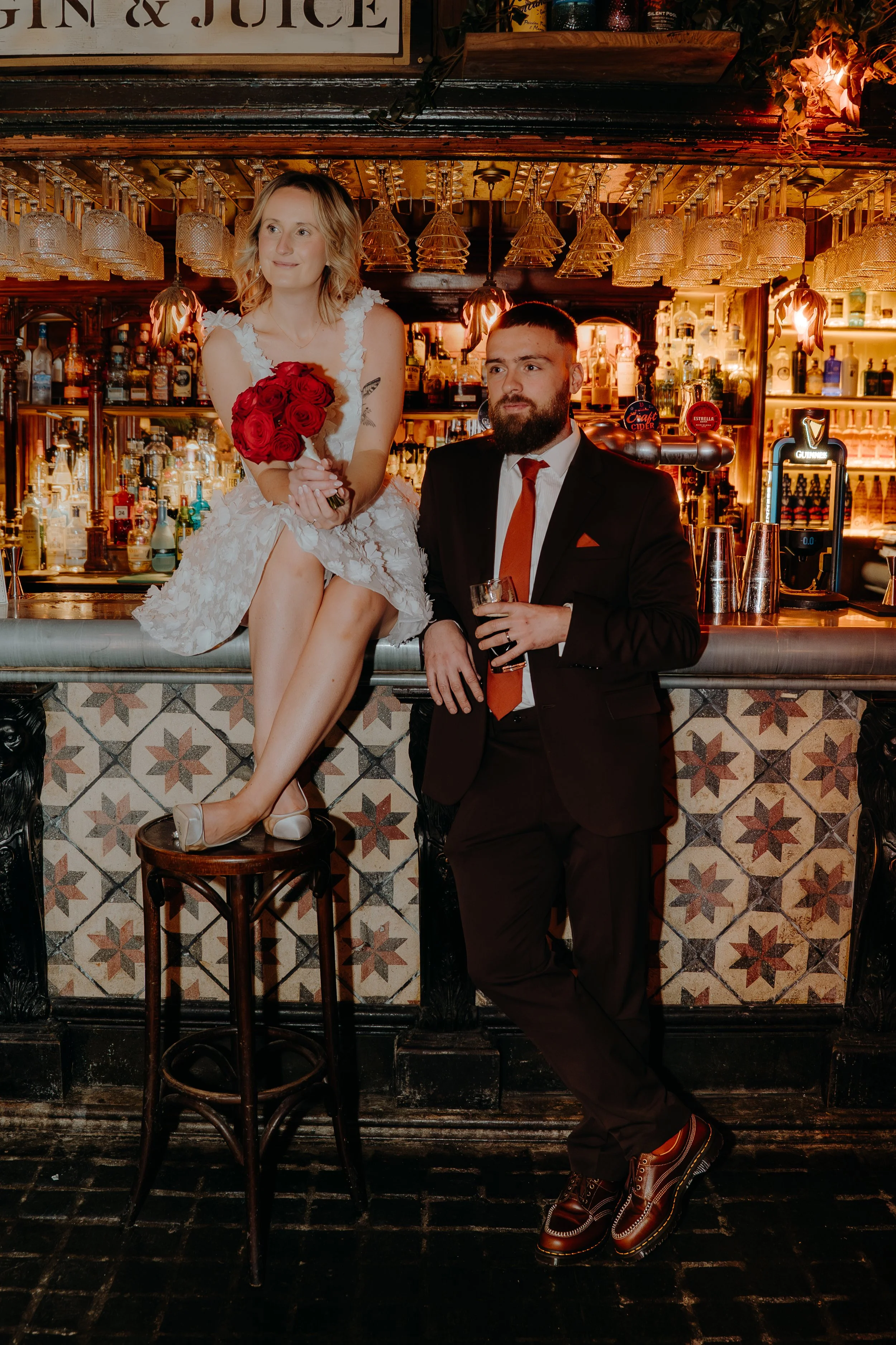 A woman with blonde wavy hair in a white dress holding a bouquet of red roses sits on a stool at a bar, with a man in a black suit and red tie leaning against the bar, holding a drink. Iris willow photography bar 131 Cheltenham