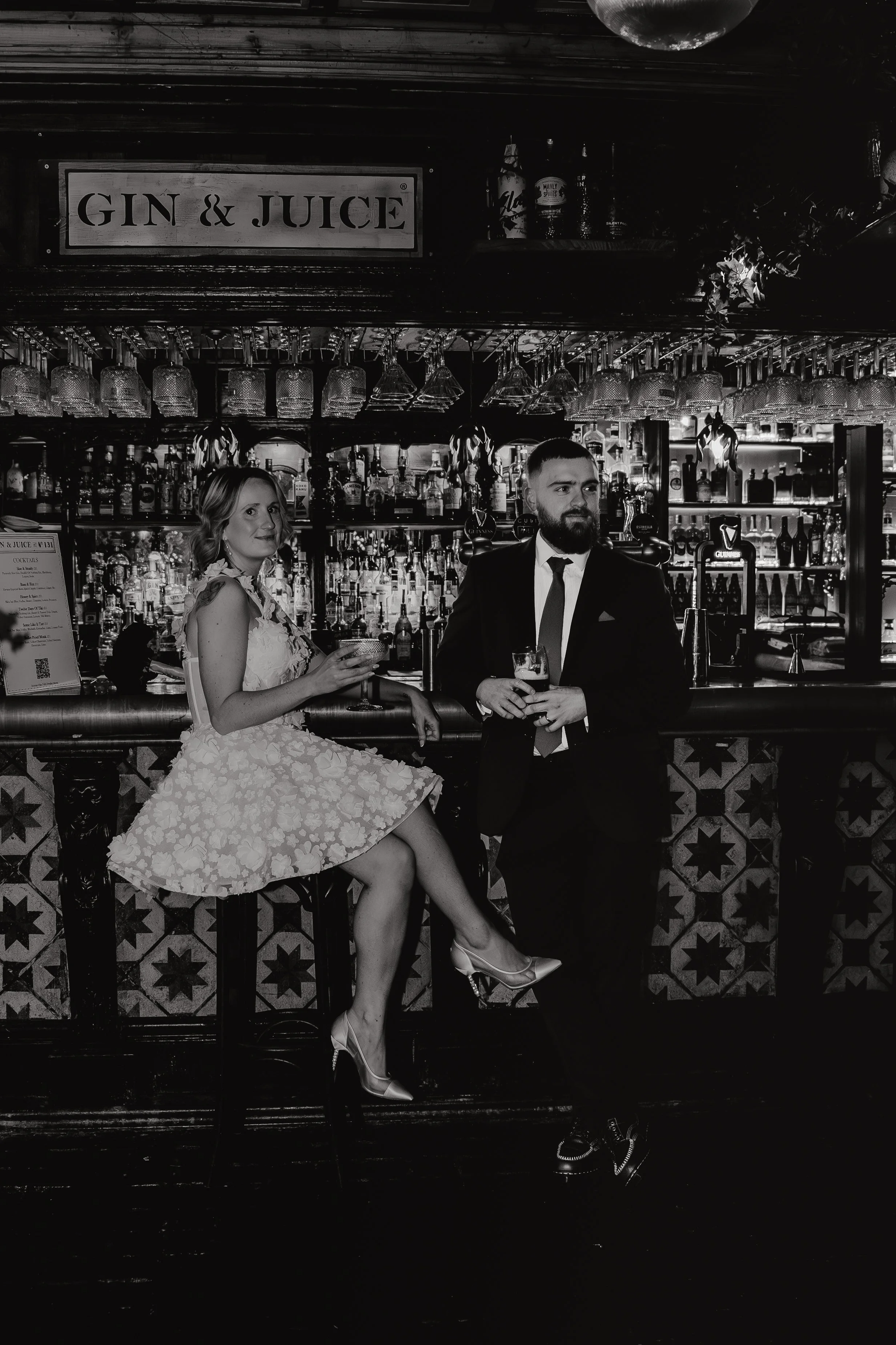 A bride sitting on a bar stool, holding a cocktail, the groom standing next to her, in bar 131 Cheltenham with bottles and glasses behind the counter, and a sign that reads 'GIN & JUICE' hanging above Iris willow photography 