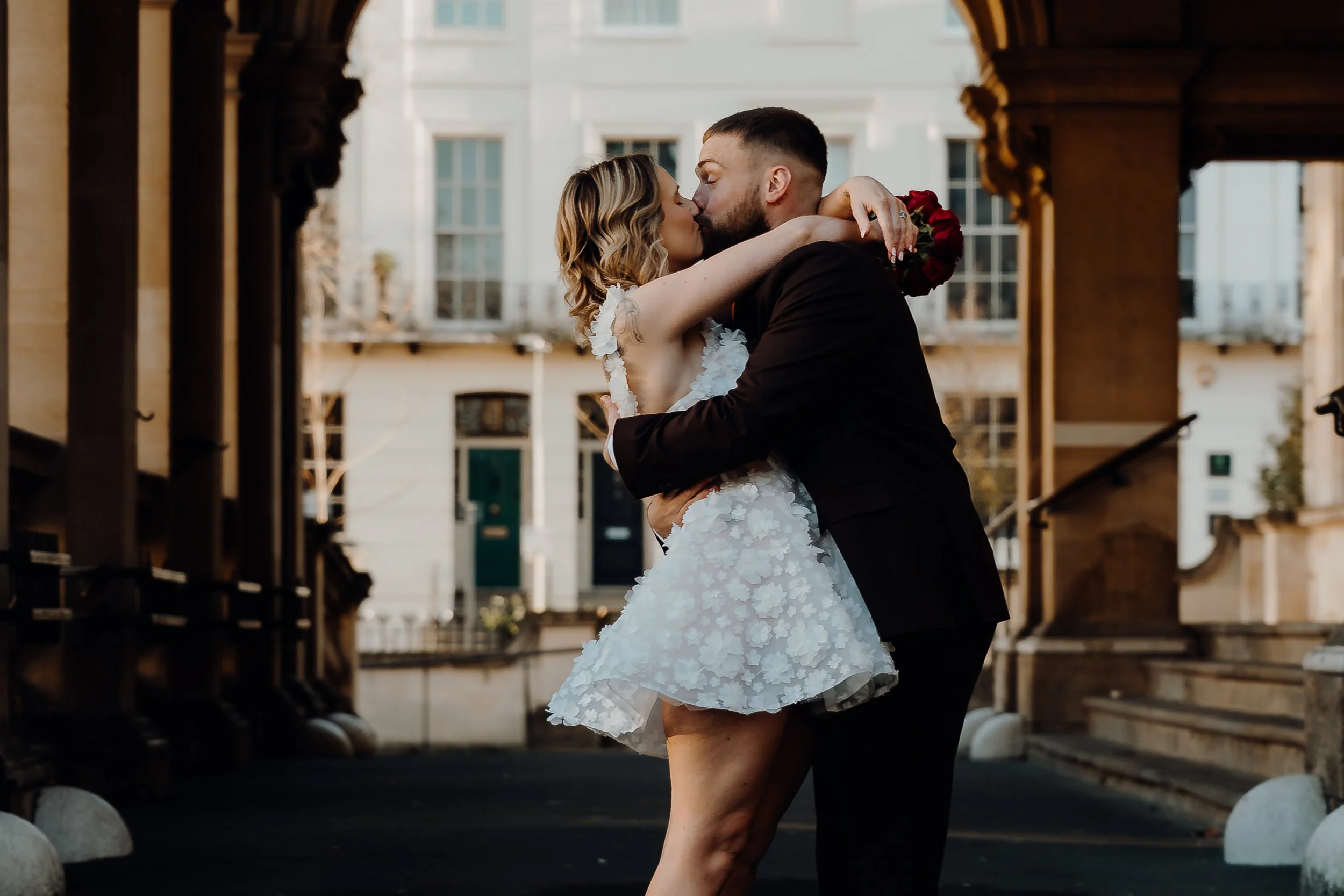 A couple kissing outdoors, woman in a white floral lace wedding dress with floral detailing and man in a black suit, with city buildings in the background.