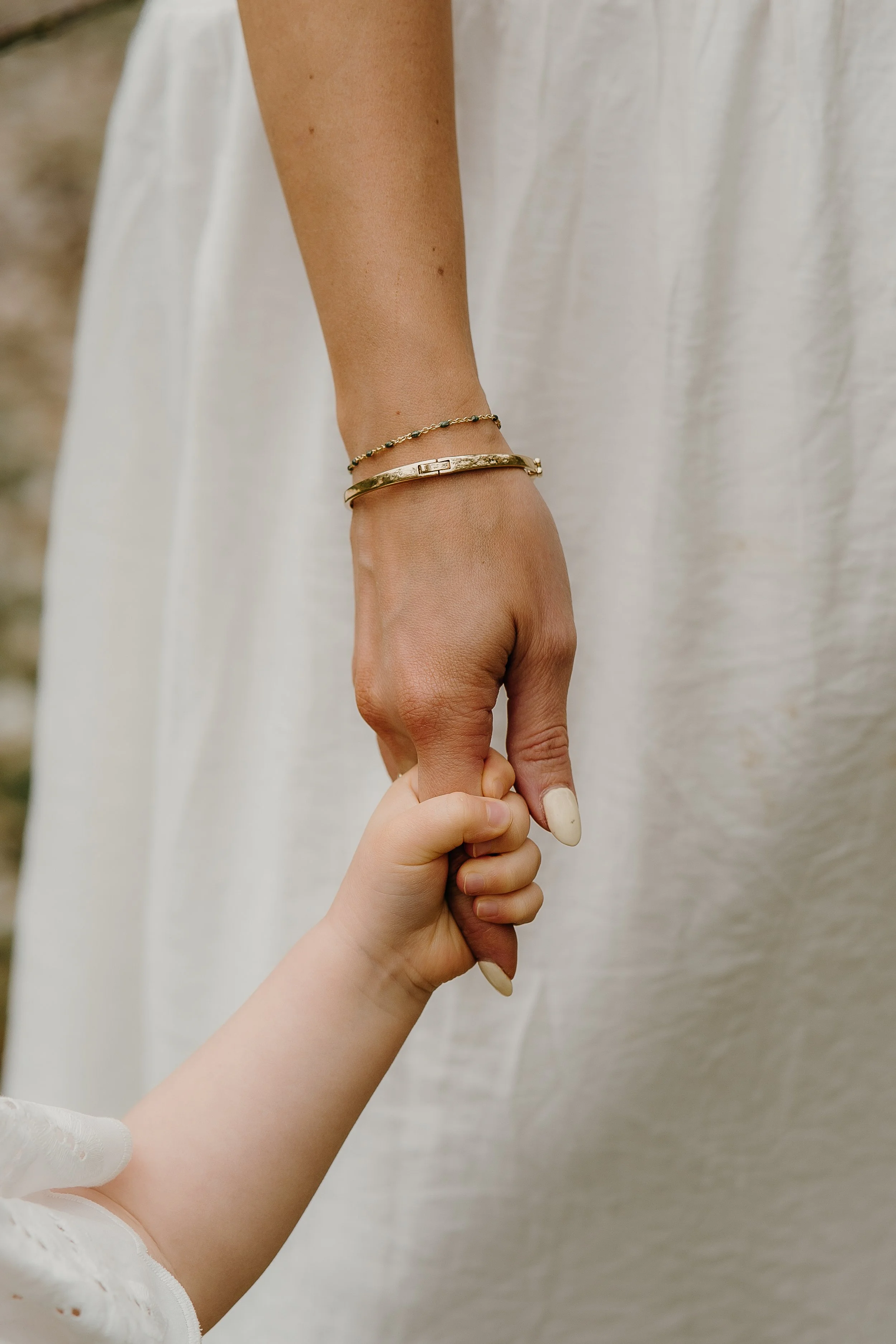 Close-up of a child's hand holding an adult woman's hand, with both wearing jewelry, against a blurred light-colored background.