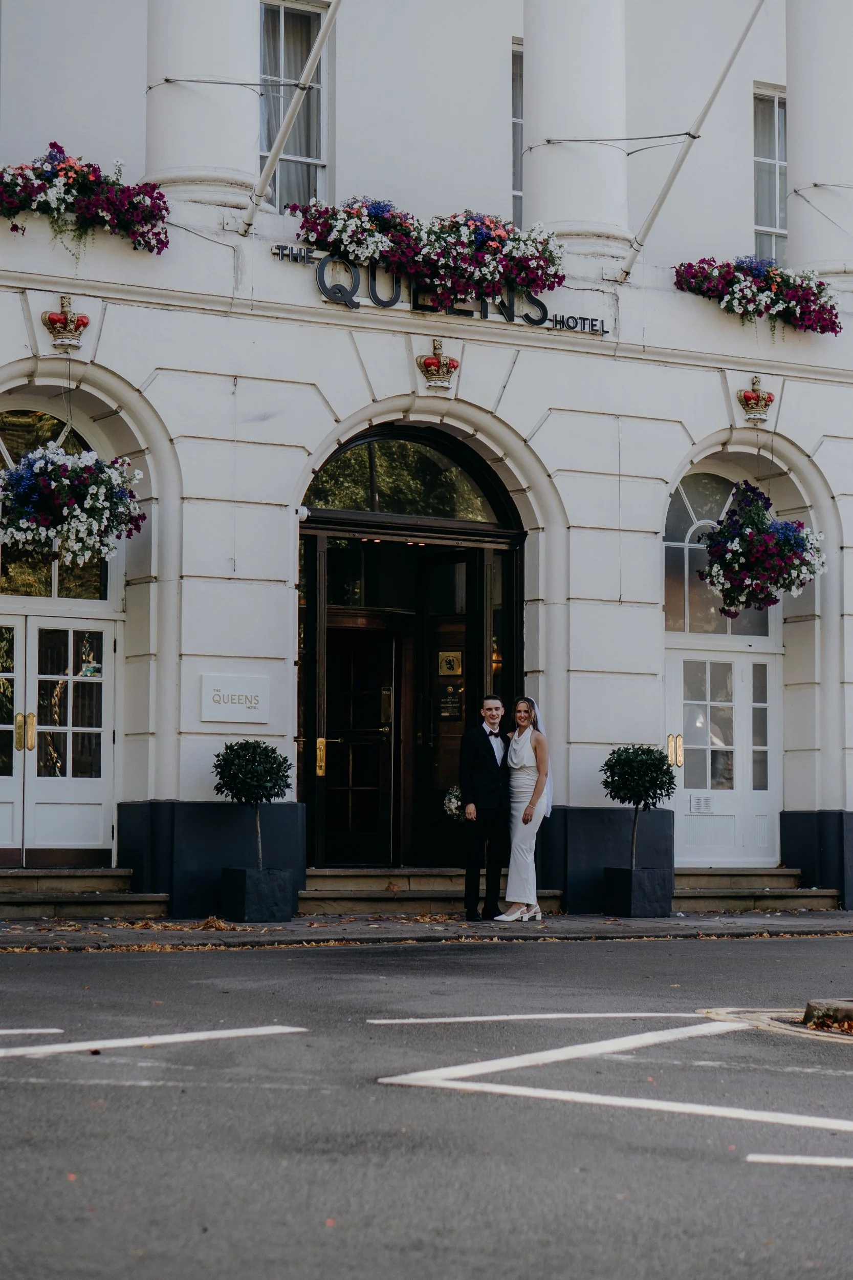 A man and a woman dressed in formal attire stand outside the Queen's Hotel, located in a white building with flowers and decorative crowns above the windows. Iris willow photography bar 131 Cheltenham 