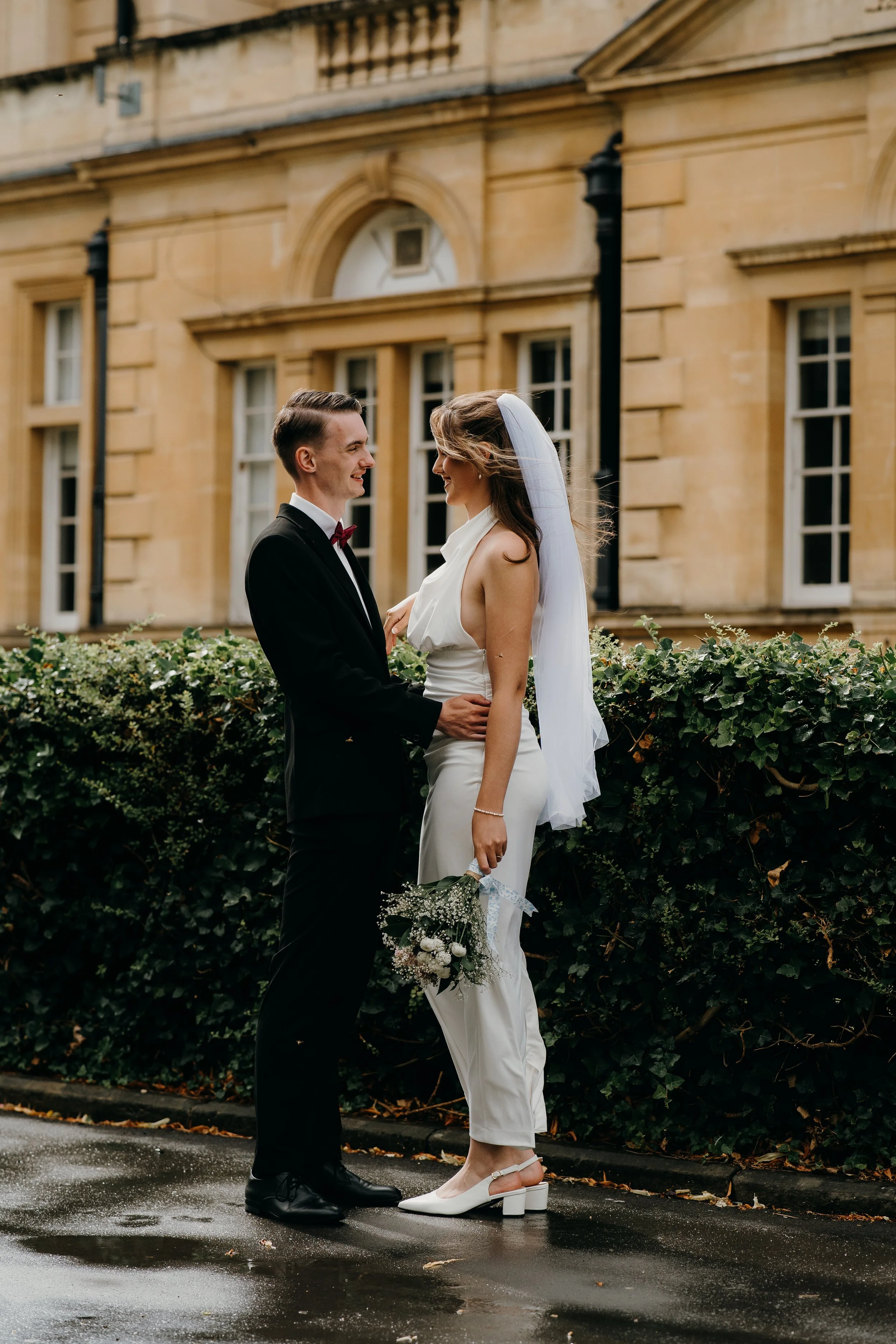 A bride and groom standing face to face outdoors, holding hands and smiling at each other on a wet pavement in front of a historic building with large windows and ivy. Iris willow photography bar 131 Cheltenham