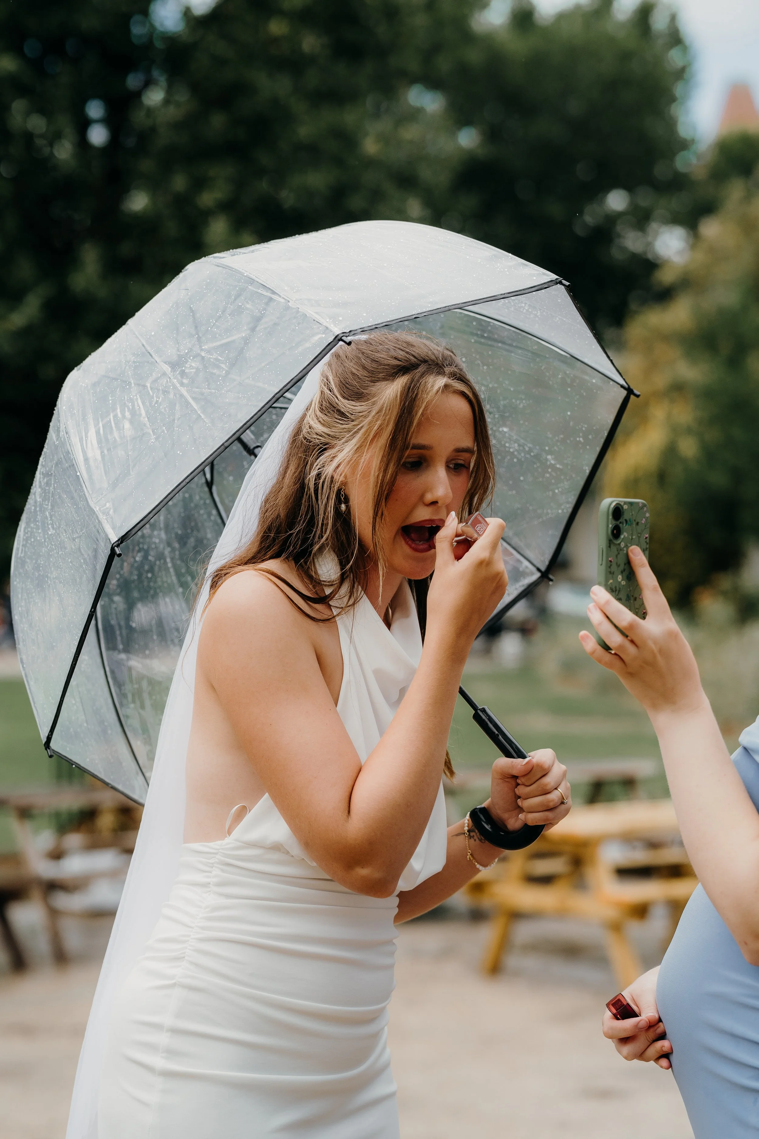 A woman in a white dress is applying lipstick under an umbrella on a rainy day outdoors.