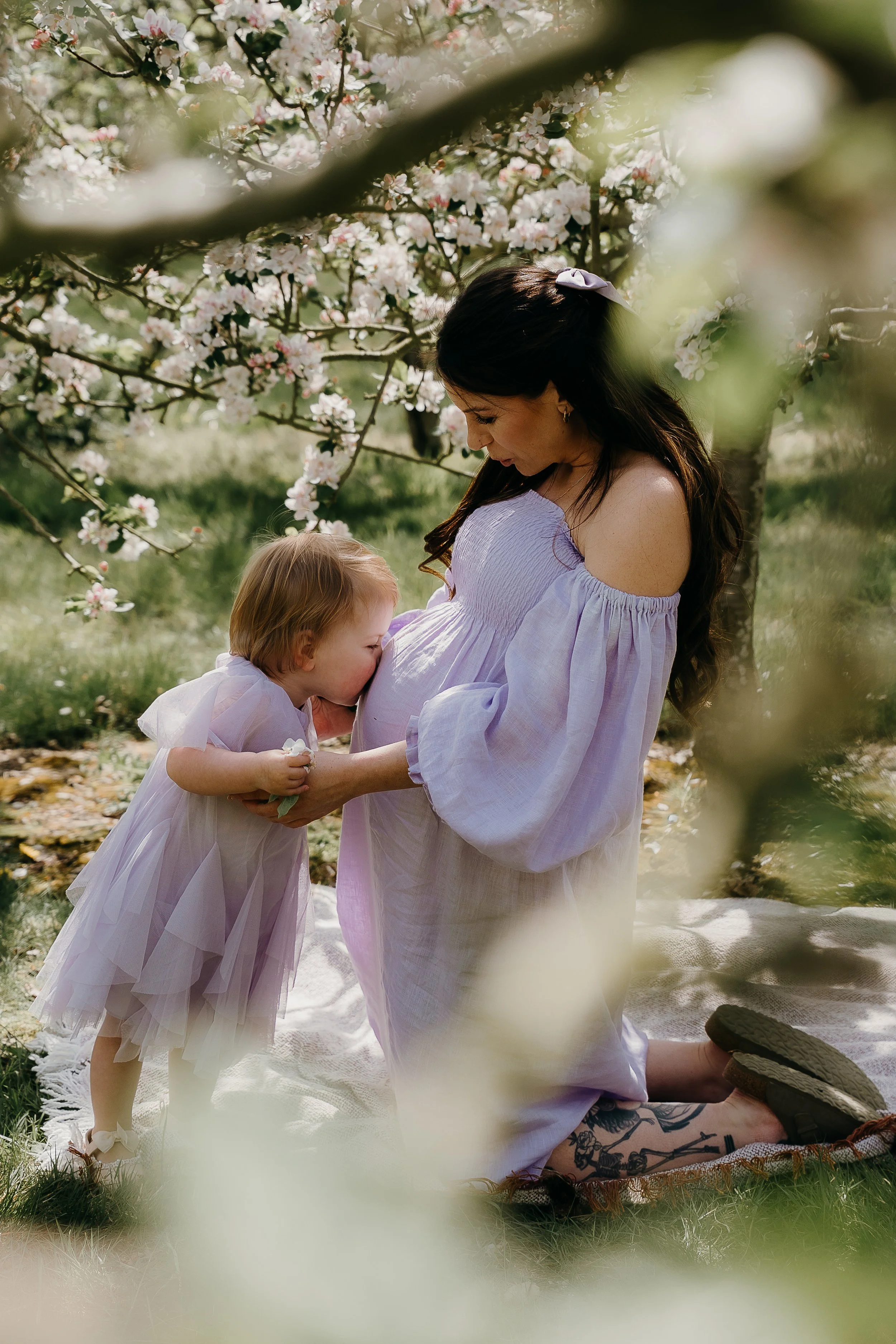 A woman in a white dress with off-the-shoulder sleeves kneeling on a blanket in a garden, breastfeeding a young child dressed in a pink tutu, under a blooming tree with pink and white flowers.